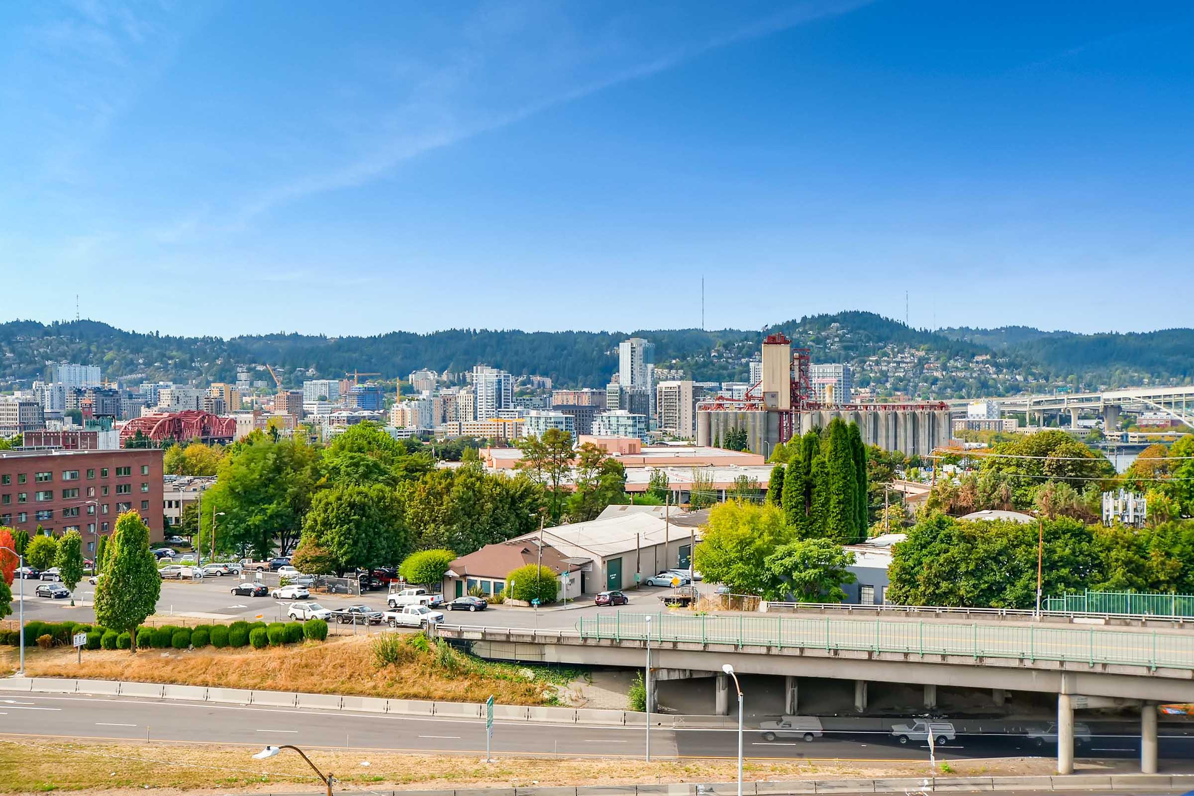A panoramic view of a city skyline with tall buildings and greenery in the foreground. The scene includes highways and a river, set against a backdrop of mountains under a clear blue sky. The landscape showcases a blend of urban and natural elements, highlighting the vibrancy of city life.