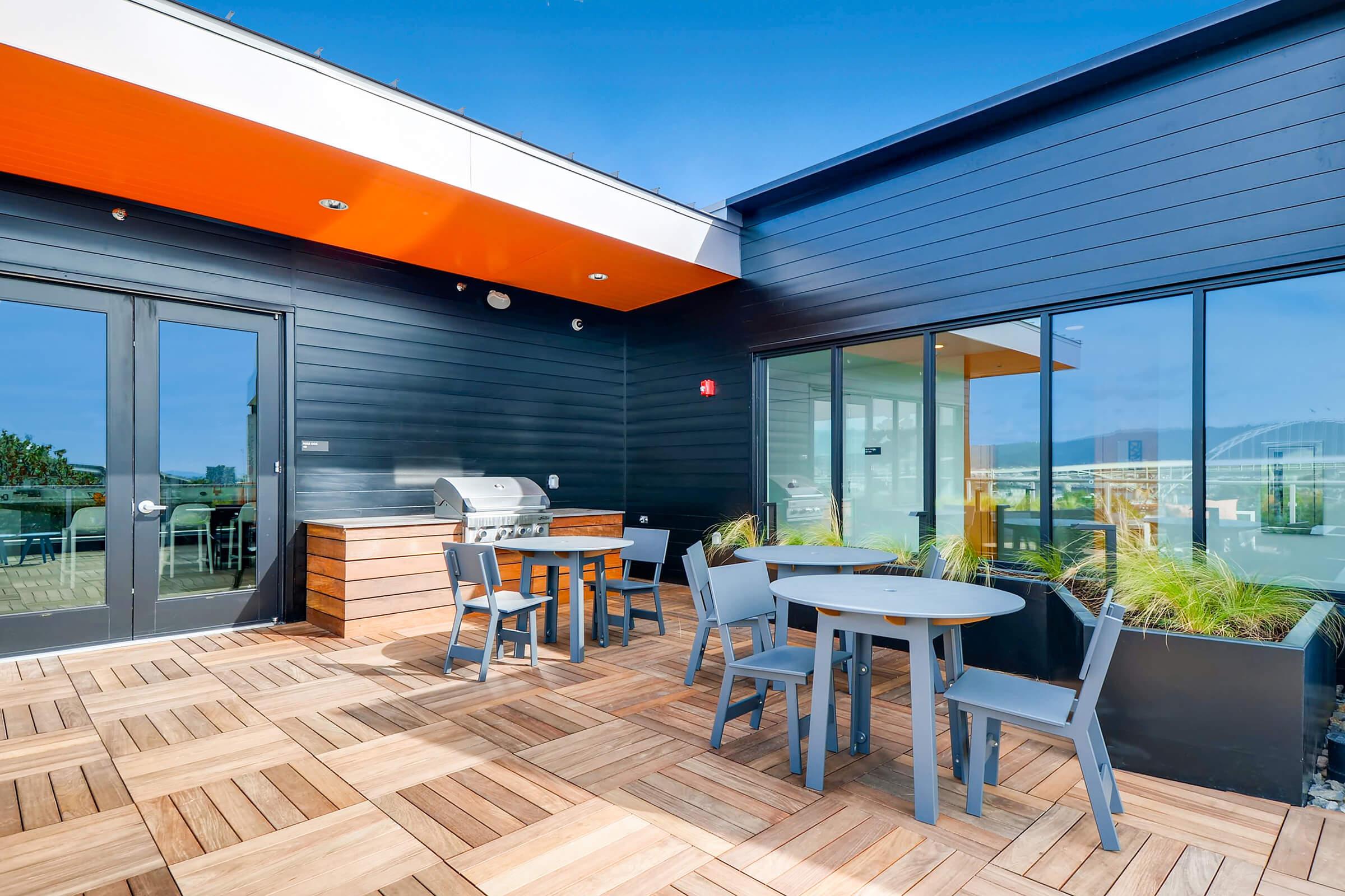 A modern rooftop terrace featuring a wooden deck, outdoor dining table with chairs, a grill, and large windows. The space is well-lit with a blue sky in the background, showcasing a stylish and comfortable area for outdoor gatherings.
