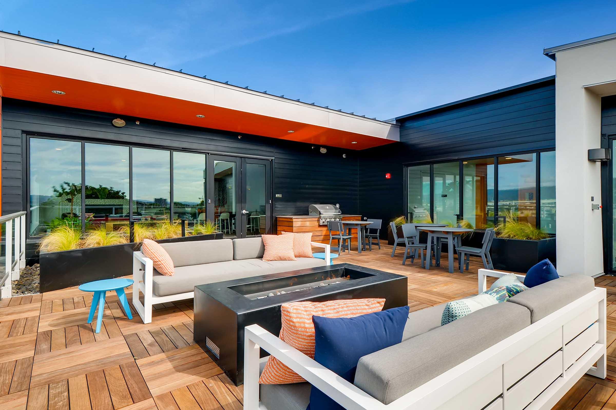 Modern rooftop patio featuring stylish outdoor furniture, including gray sofas with colorful cushions, a glass fire pit, and a small blue side table. The space is surrounded by greenery and large windows, with a barbecue grill and dining table visible in the background beneath a clear blue sky.