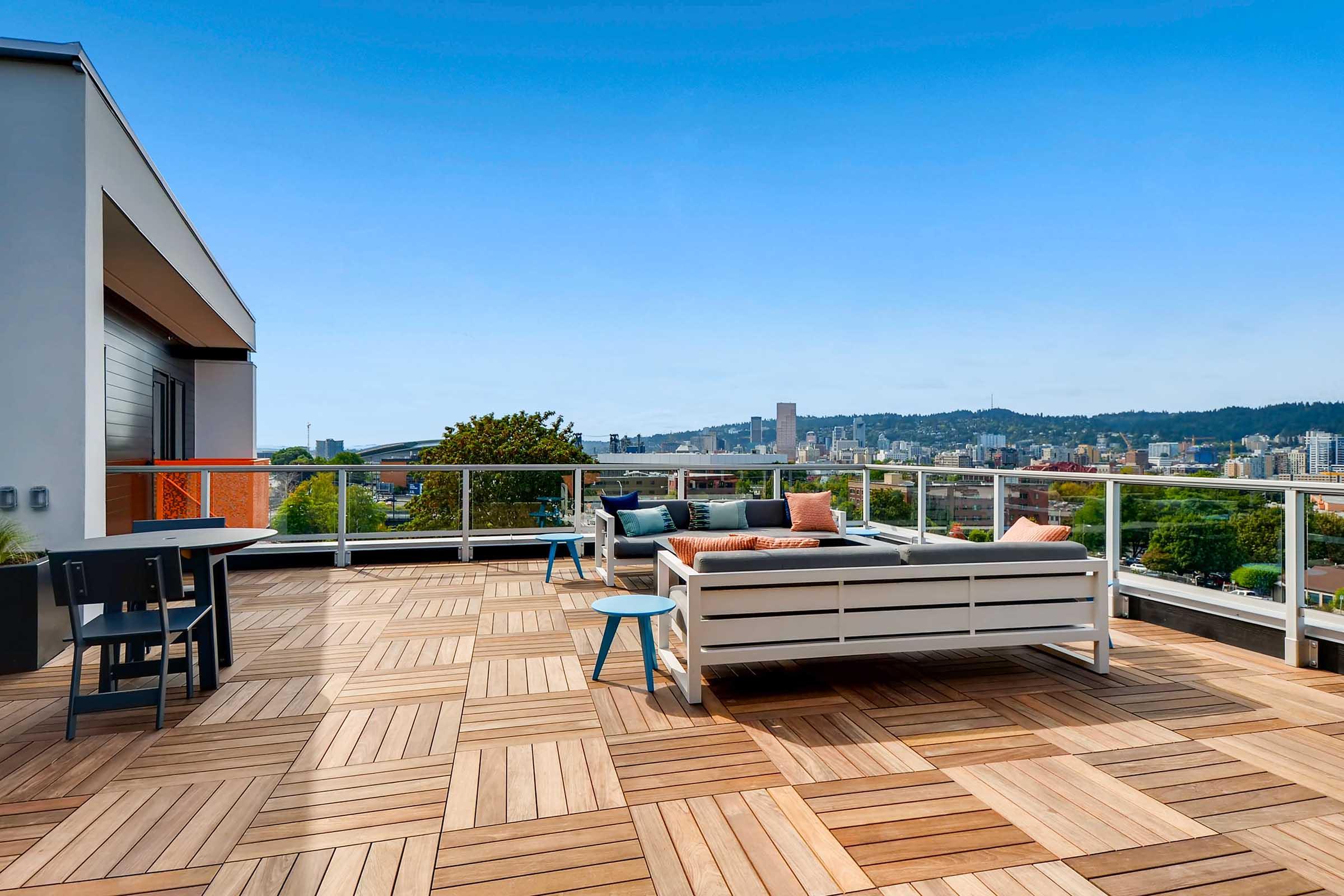 A modern rooftop terrace featuring a wooden deck, stylish seating area with cushions, and a small table. The space offers panoramic views of a city skyline under a clear blue sky, surrounded by greenery and urban architecture.