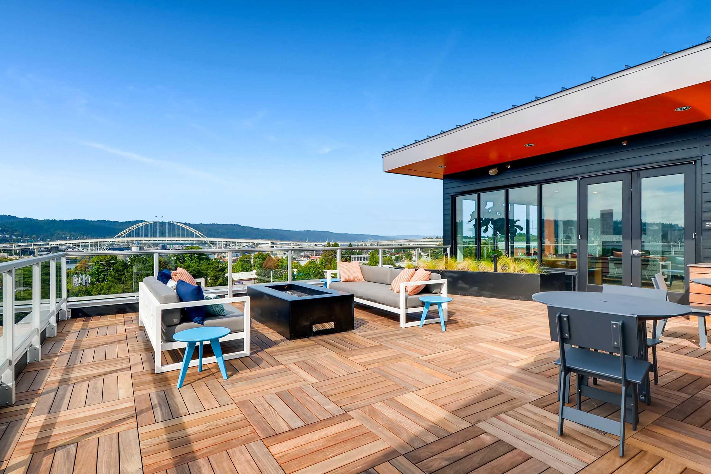A spacious rooftop terrace with wooden flooring, featuring modern outdoor furniture including a sectional sofa, coffee table, and dining table. The backdrop includes a scenic view of a bridge and green hills under a bright blue sky.