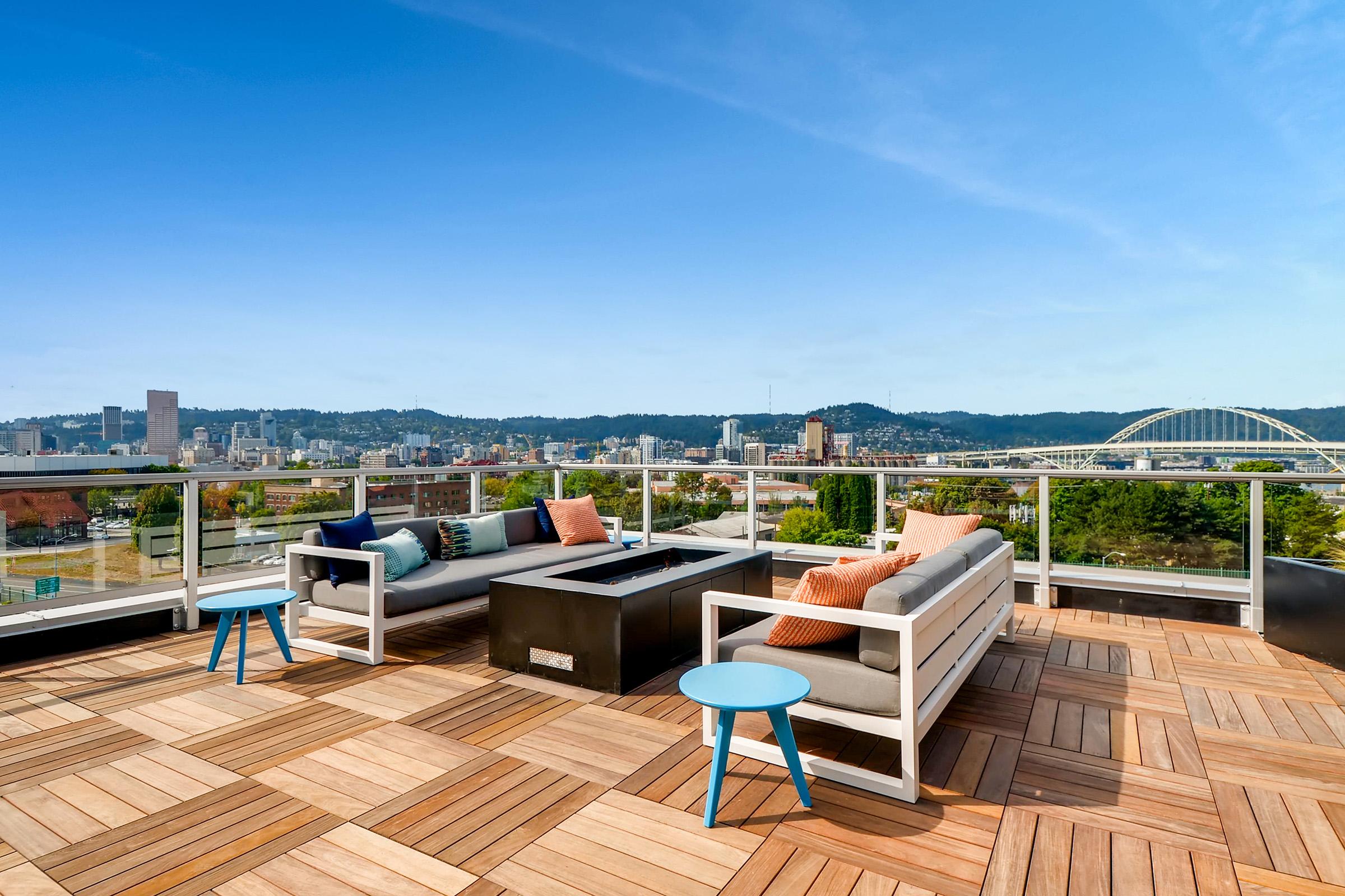 A rooftop terrace featuring comfortable seating with cushions, a small table, and a fire pit. The space is surrounded by glass railings, offering a panoramic view of a city skyline and distant hills under a clear blue sky. The wooden deck adds a cozy, modern touch to the outdoor area.