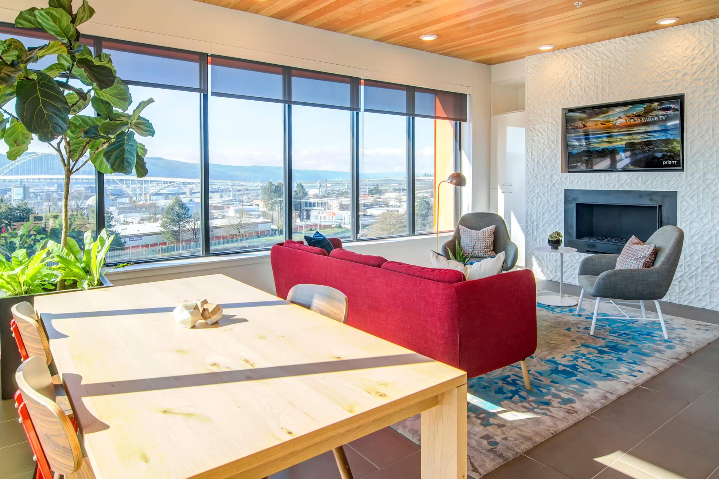 Bright, modern living space featuring a red sofa, two gray armchairs, and a dining table. Large windows offer a view of a cityscape with a bridge in the background, while indoor plants enhance the decor. The room has a cozy, inviting atmosphere with stylish furnishings and natural light.