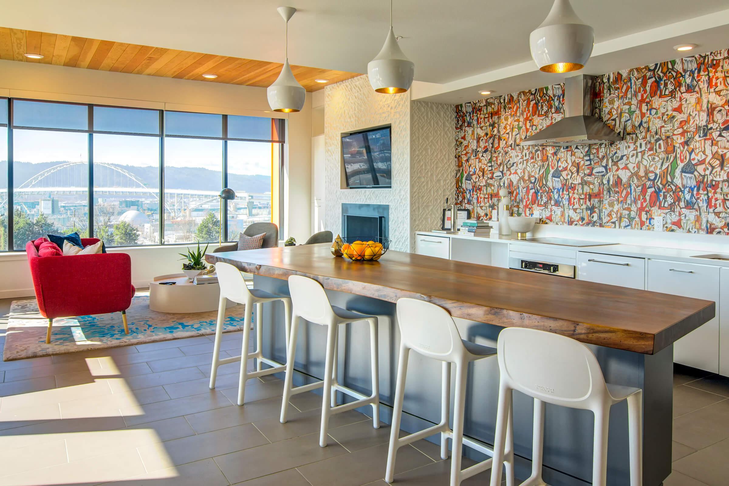 A modern kitchen with a large wooden island and white bar stools. Large windows provide a view of a bridge and mountains. A red chair and a colorful wall art feature add brightness, while the kitchen is equipped with sleek appliances and a cozy fireplace. Fresh fruit sits in a bowl on the island.