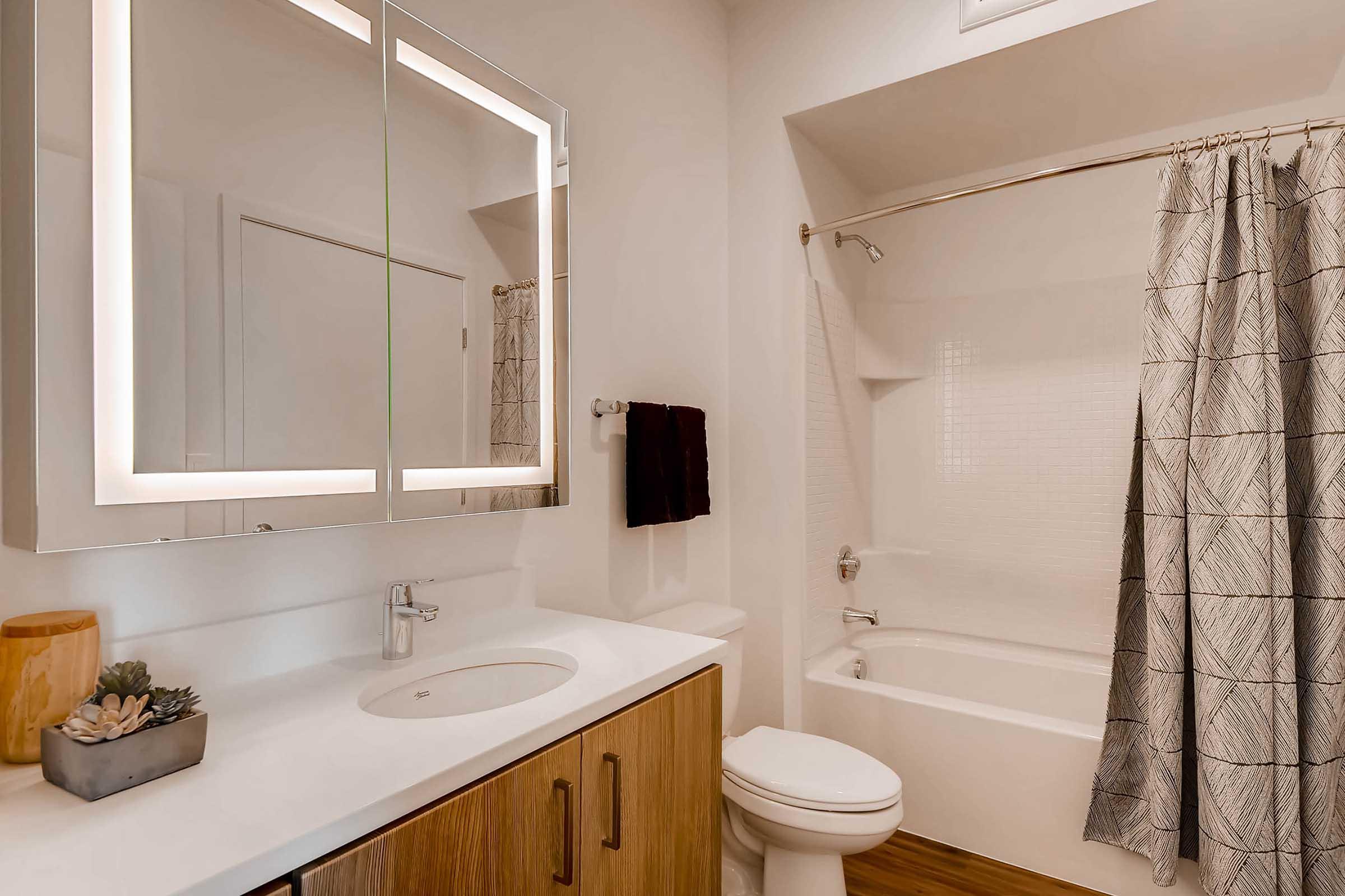 A modern bathroom featuring a white countertop with a sink, a large illuminated mirror, a bathtub with a shower curtain, and a wooden vanity. The walls are light-colored, complemented by a soft gray shower curtain and a small decorative plant on the counter. A towel is hung on the side.