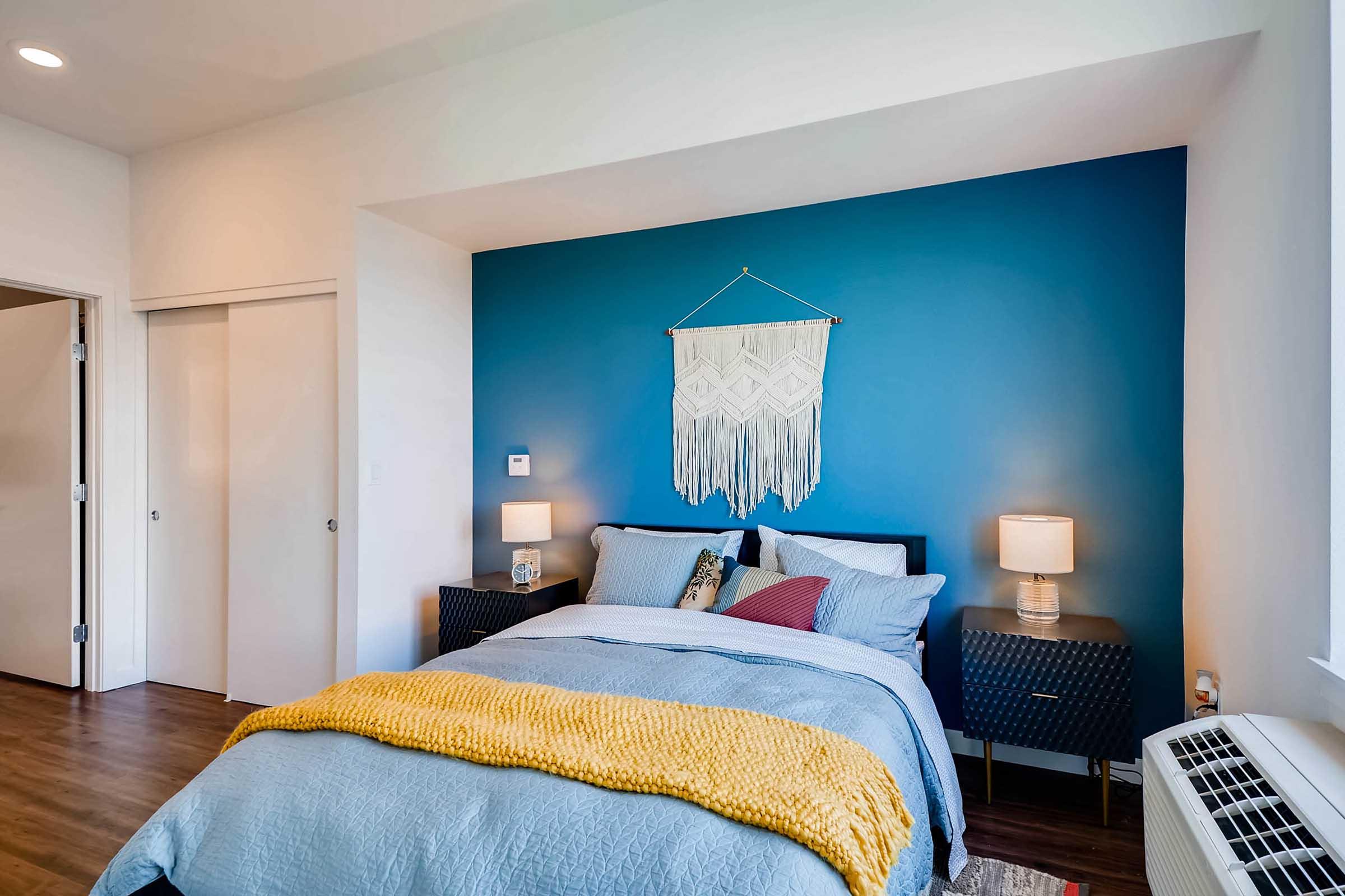 A cozy bedroom featuring a blue accent wall, a neatly made bed with blue and yellow bedding, two bedside tables with lamps, and a macramé wall hanging. There are large windows allowing natural light, and light wooden flooring adds warmth to the space.