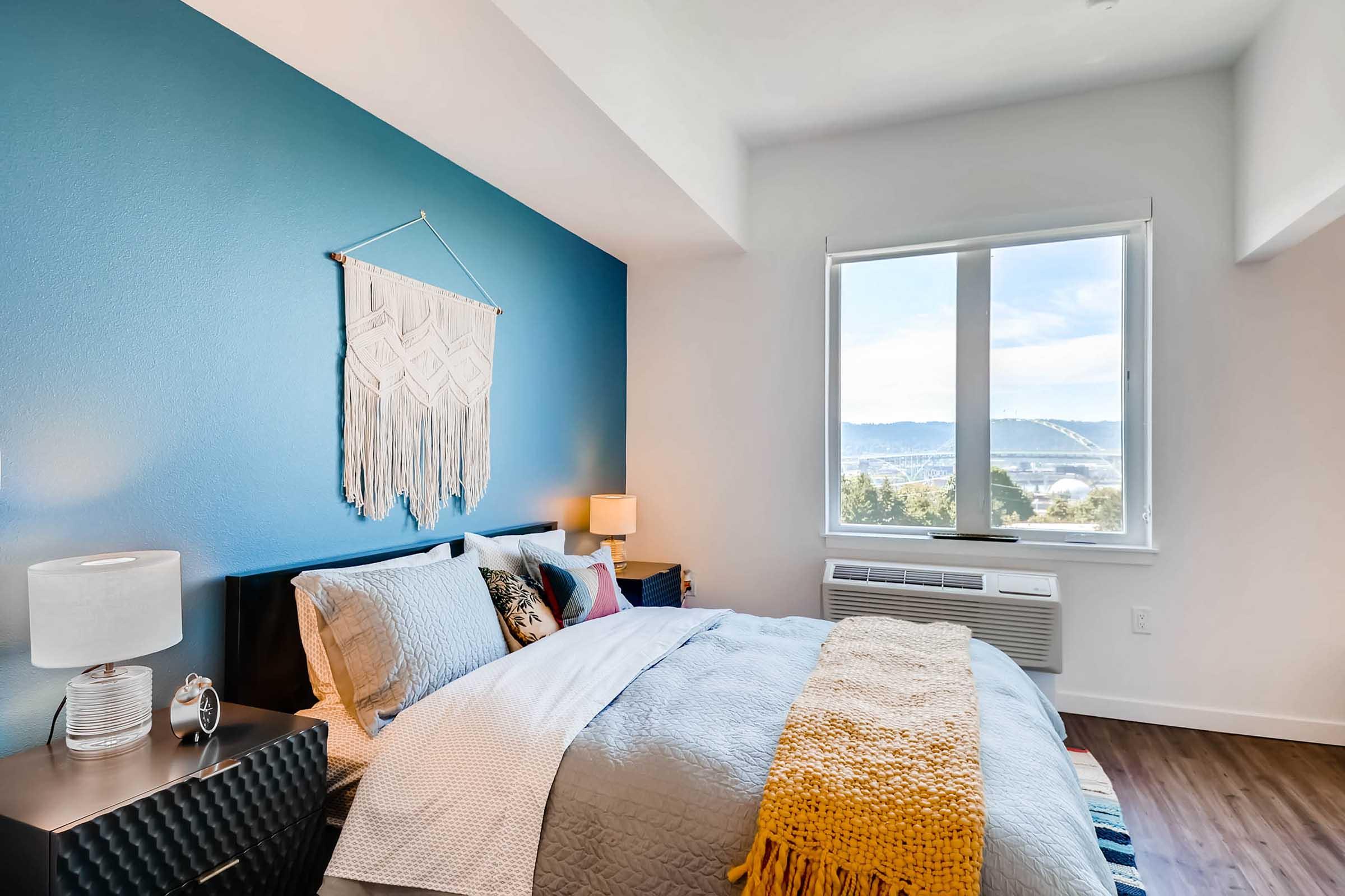 A cozy bedroom featuring a bed with colorful bedding, decorative pillows, and a knitted throw. A nightstand with a lamp is positioned beside the bed. The wall behind the bed is painted in a deep blue color, adorned with a macramé wall hanging. A window provides natural light and offers a view of the outdoors.