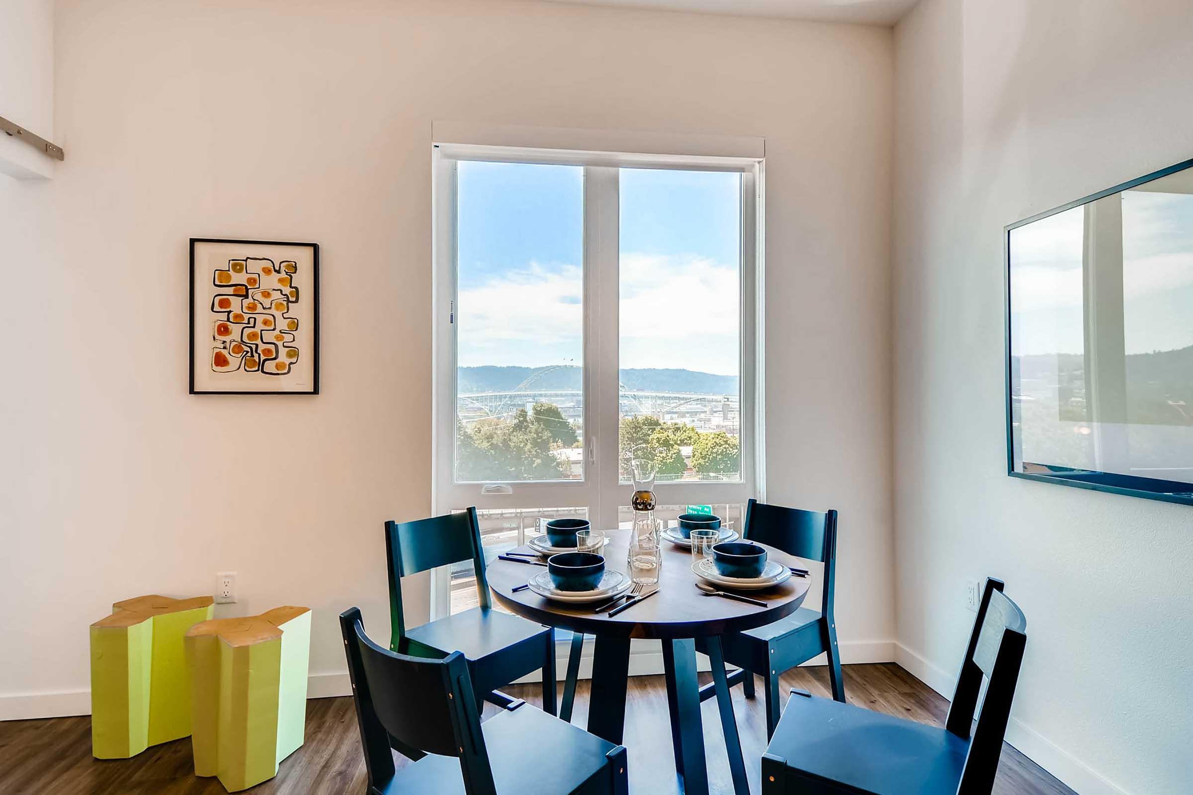 A modern dining area featuring a round table set for four with blue plates and glasses. Bright natural light flows through large windows, offering a scenic view of the outdoors. A colorful artwork is hung on the wall, and two unique stools are placed nearby, adding a contemporary touch to the space.