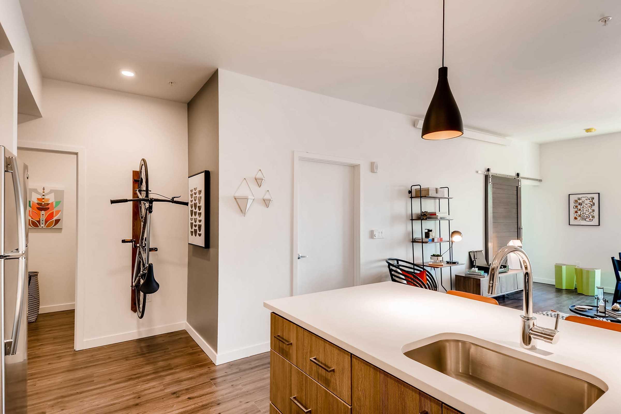Modern open-concept kitchen with a central island and wooden cabinetry. A bicycle is mounted on the wall near a doorway leading to a hallway. The space features decorative wall art, a hanging pendant light, and a living area visible in the background with colorful furniture and shelving.