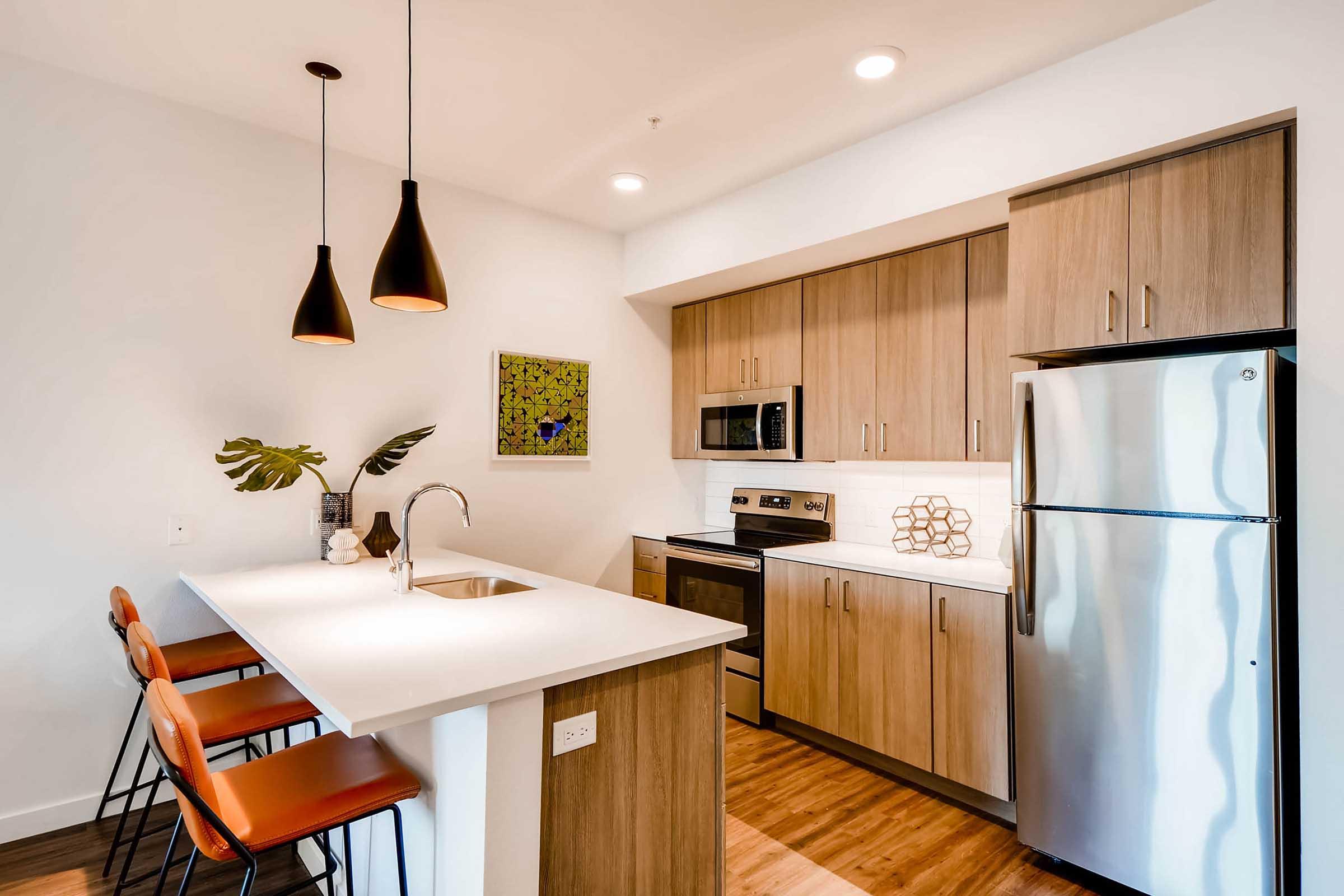 Modern kitchen featuring wooden cabinets, stainless steel appliances, and a white countertop. There are three orange bar stools at the kitchen island, and pendant lighting hangs above. A piece of abstract art is displayed on the wall, adding a pop of color to the space.