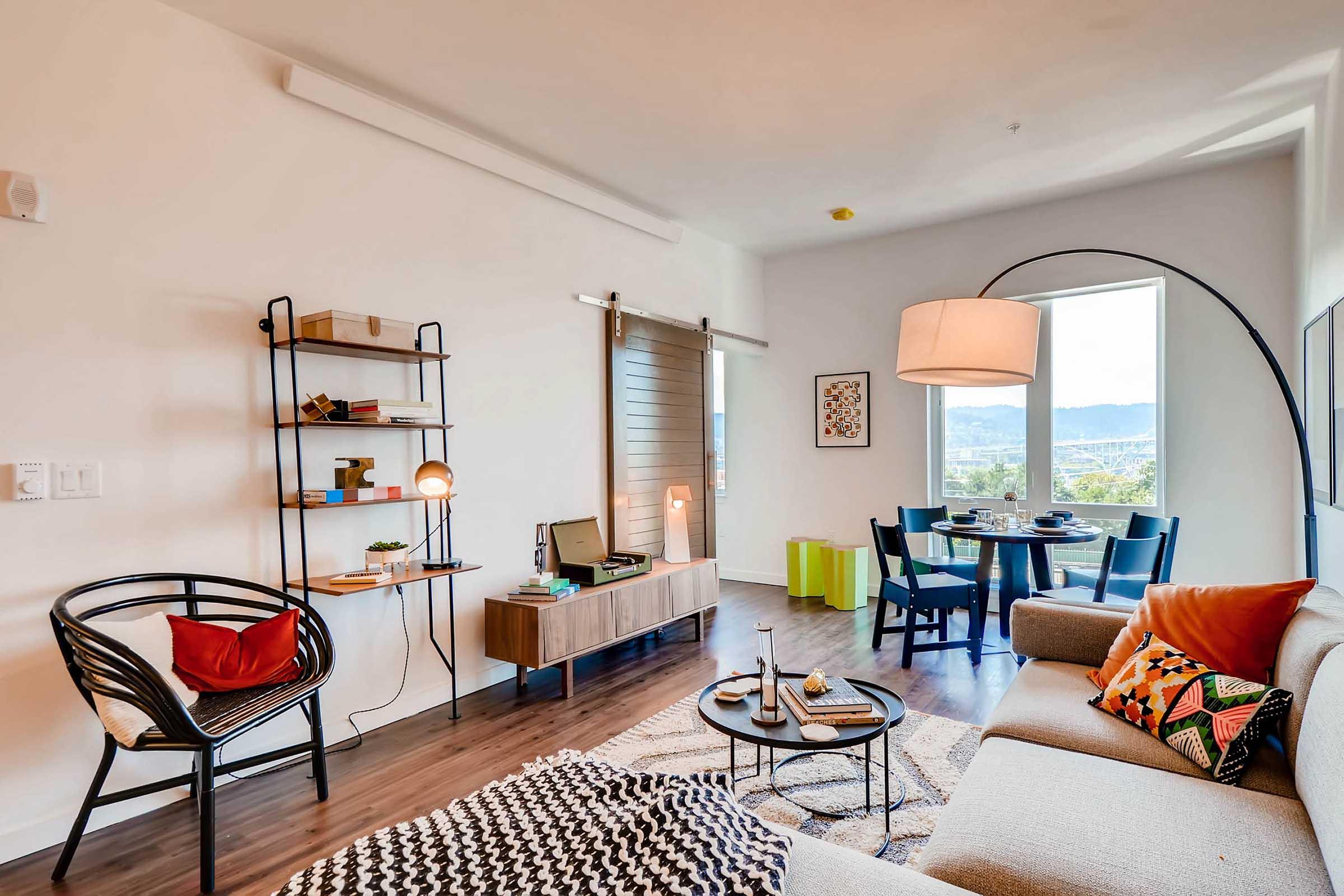 A modern living room featuring a cozy sofa adorned with colorful cushions, a stylish armchair, and a coffee table with decorative items. A bookshelf and lamp add to the ambiance, while a dining table for four is visible nearby. Large windows provide natural light and a view of the outdoors.