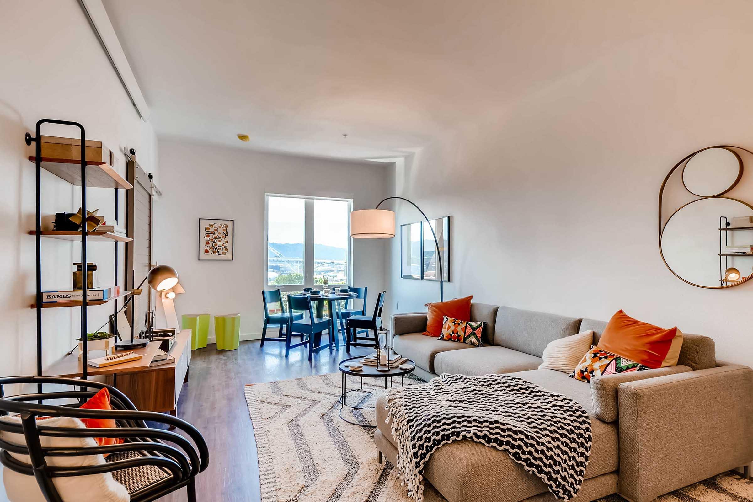 A modern living room featuring a light-colored sofa with colorful throw pillows, a cozy blanket draped over one side, and a round coffee table. There's a stylish floor lamp, decorative shelves, and artwork on the walls. A dining area with a round table and chairs is visible in the background, along with large windows offering a view.