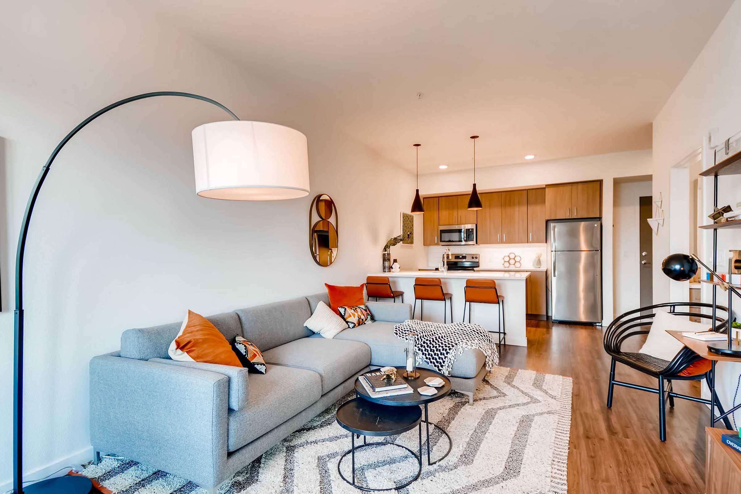 A modern living room featuring a gray sofa with decorative pillows, a round coffee table with books and a decorative item, and a stylish floor lamp. The open kitchen area has wooden cabinets, stainless steel appliances, and barstool seating. The decor includes a patterned rug and wall art, creating a cozy atmosphere.
