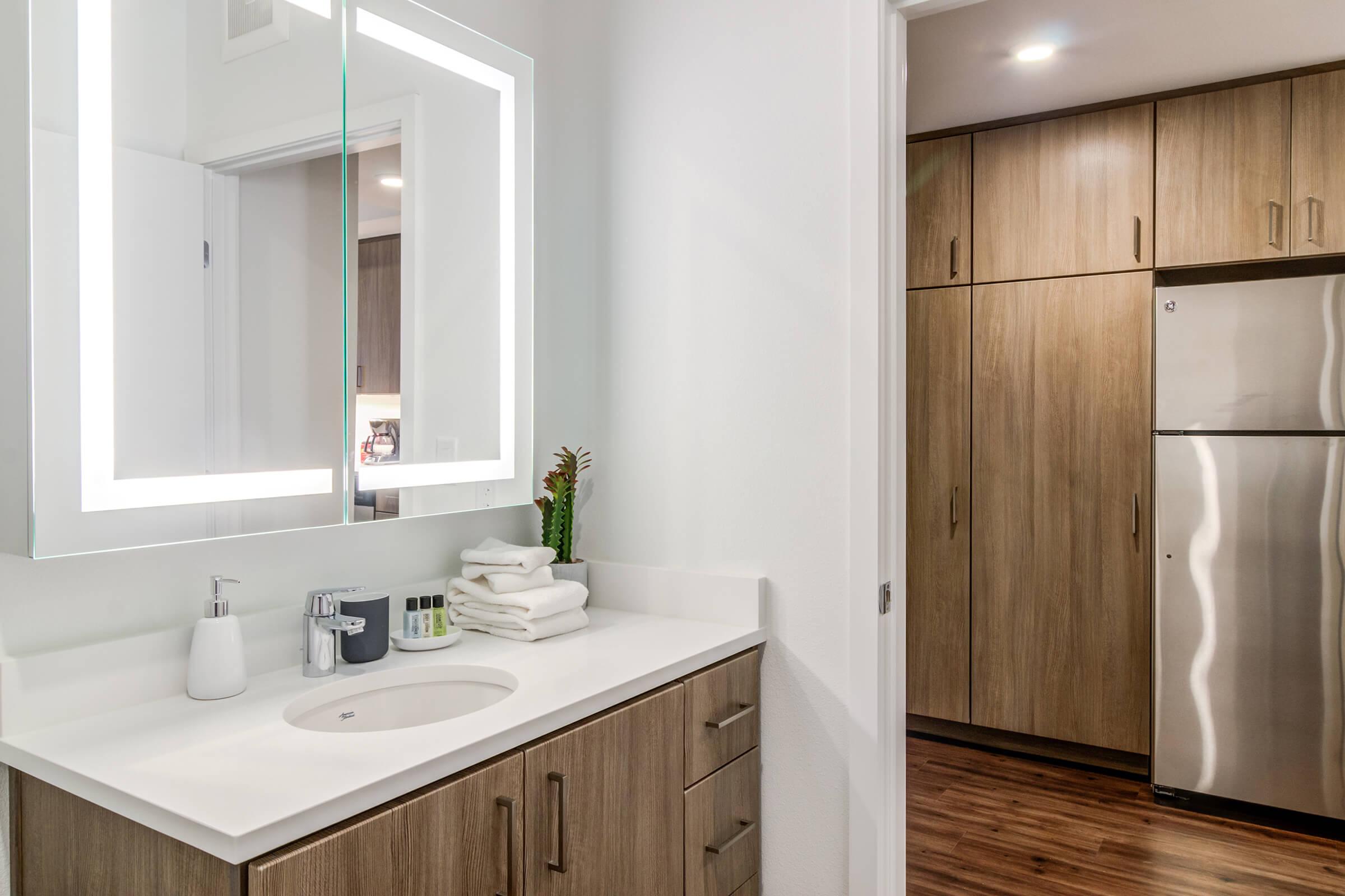 A modern bathroom featuring a sleek mirror with built-in lighting above a white sink and wooden cabinetry. A small plant is placed beside neatly stacked white towels on the countertop, with an adjacent room showing kitchen cabinets and a stainless steel refrigerator in the background.