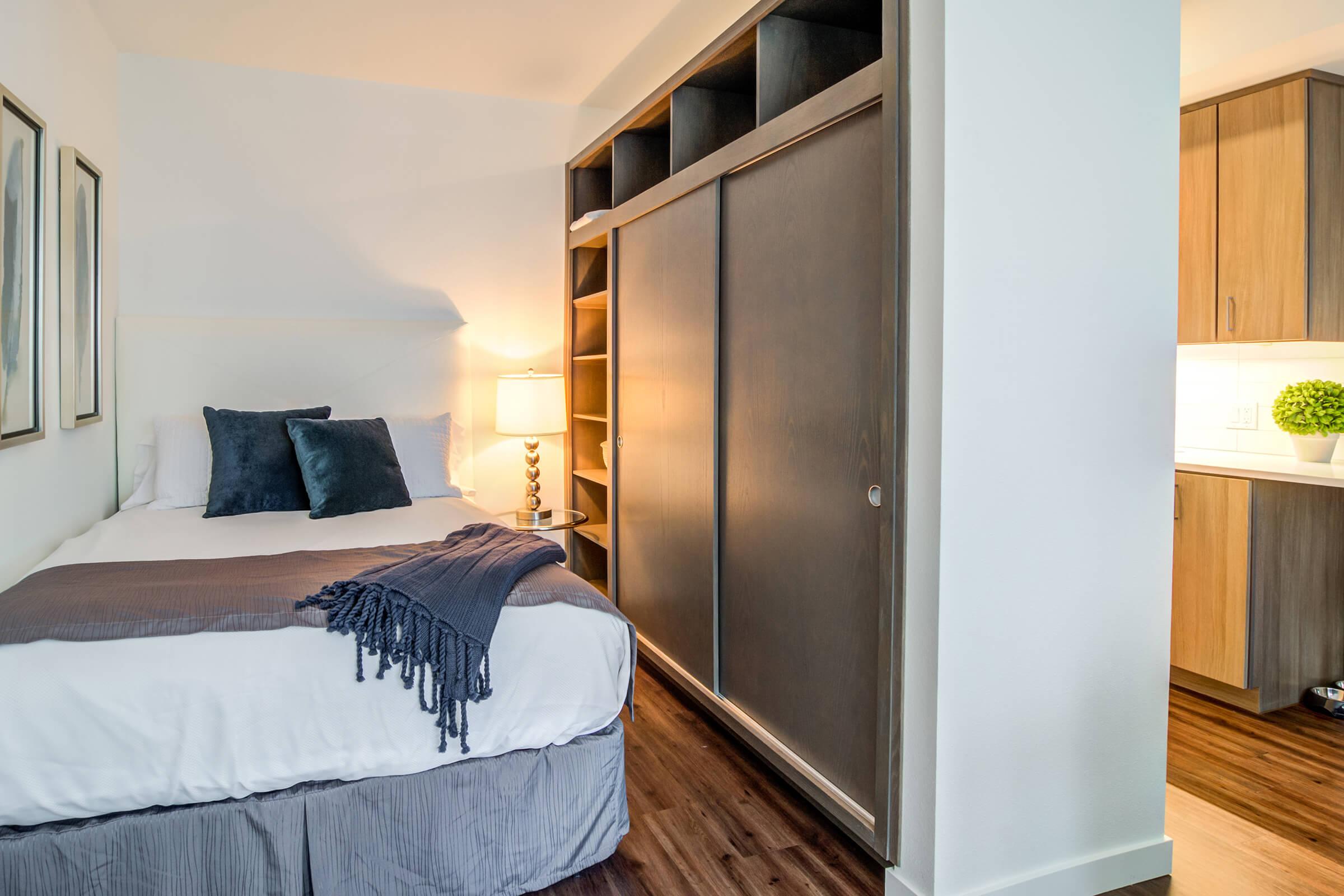 A cozy bedroom featuring a double bed with decorative pillows and a throw blanket. A bedside lamp sits on a nightstand, and a sliding wooden wardrobe is nearby. In the background, a glimpse of a kitchen can be seen, with wooden cabinetry and modern design elements. Natural light brightens the space.