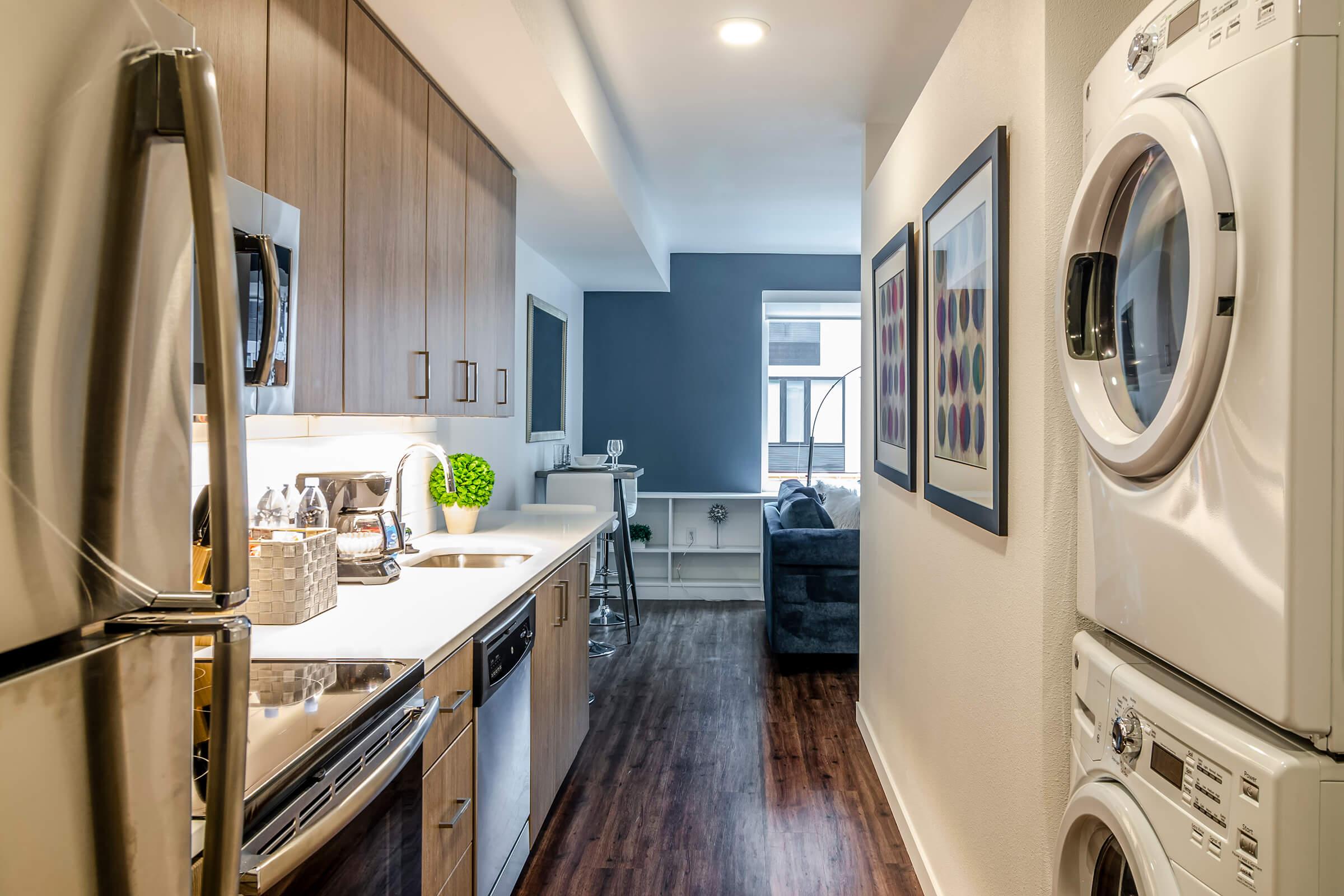 A modern kitchen with light wood cabinets and stainless steel appliances, including a refrigerator and stove. On the right, a stacked washer and dryer are visible. A small dining area with a bar stool is near a window, and a cozy living space with a blue couch is seen in the background.