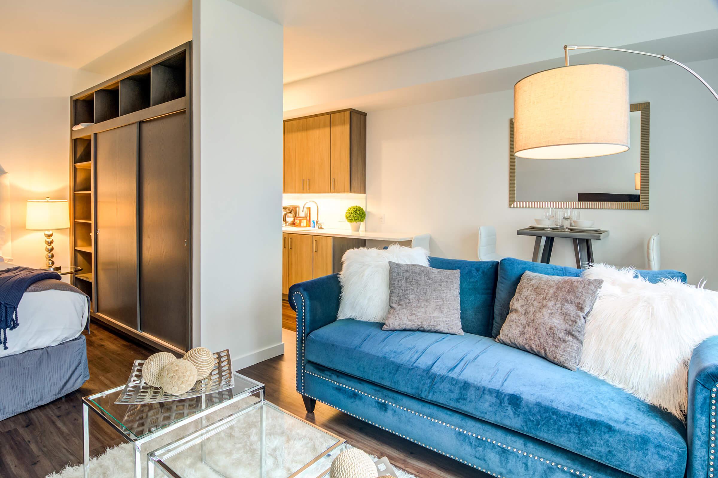 A modern living space featuring a stylish blue sofa adorned with several cozy pillows, a glass coffee table, and a soft area rug. In the background, there is a kitchen with wooden cabinets and a dining table set. The atmosphere is bright and inviting, with warm lighting and decorative accents.