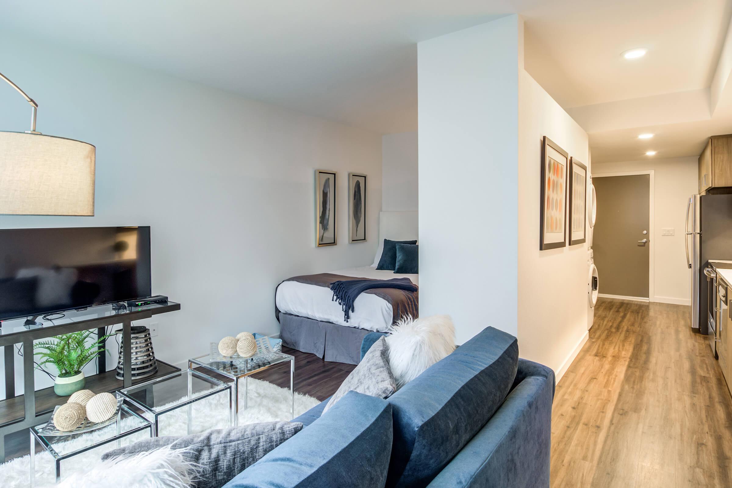 Cozy modern studio apartment featuring a living area with a blue sofa, a glass coffee table, and a television. The sleeping area is partially separated with a bed dressed in blue linens. Warm wood flooring and neutral walls create a welcoming atmosphere. Soft lighting adds to the inviting ambiance.