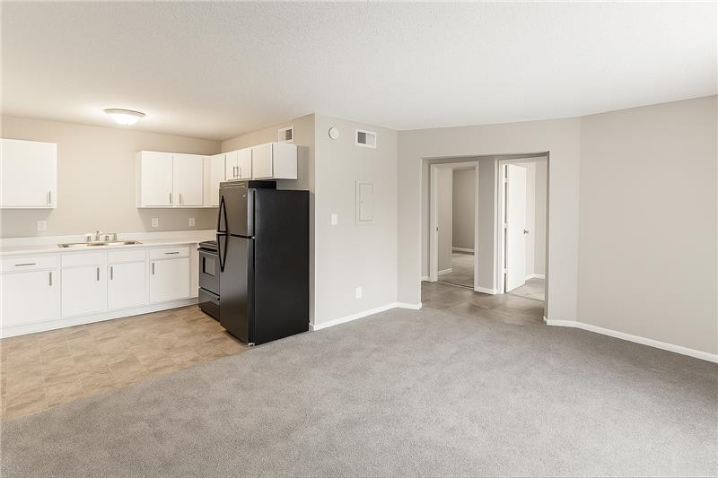 A spacious, well-lit apartment interior featuring a kitchen with white cabinets and black appliances, and a living area with beige carpet. There's a doorway leading to another room or hallway, providing additional living space. The walls are painted light gray, creating a modern and inviting atmosphere.