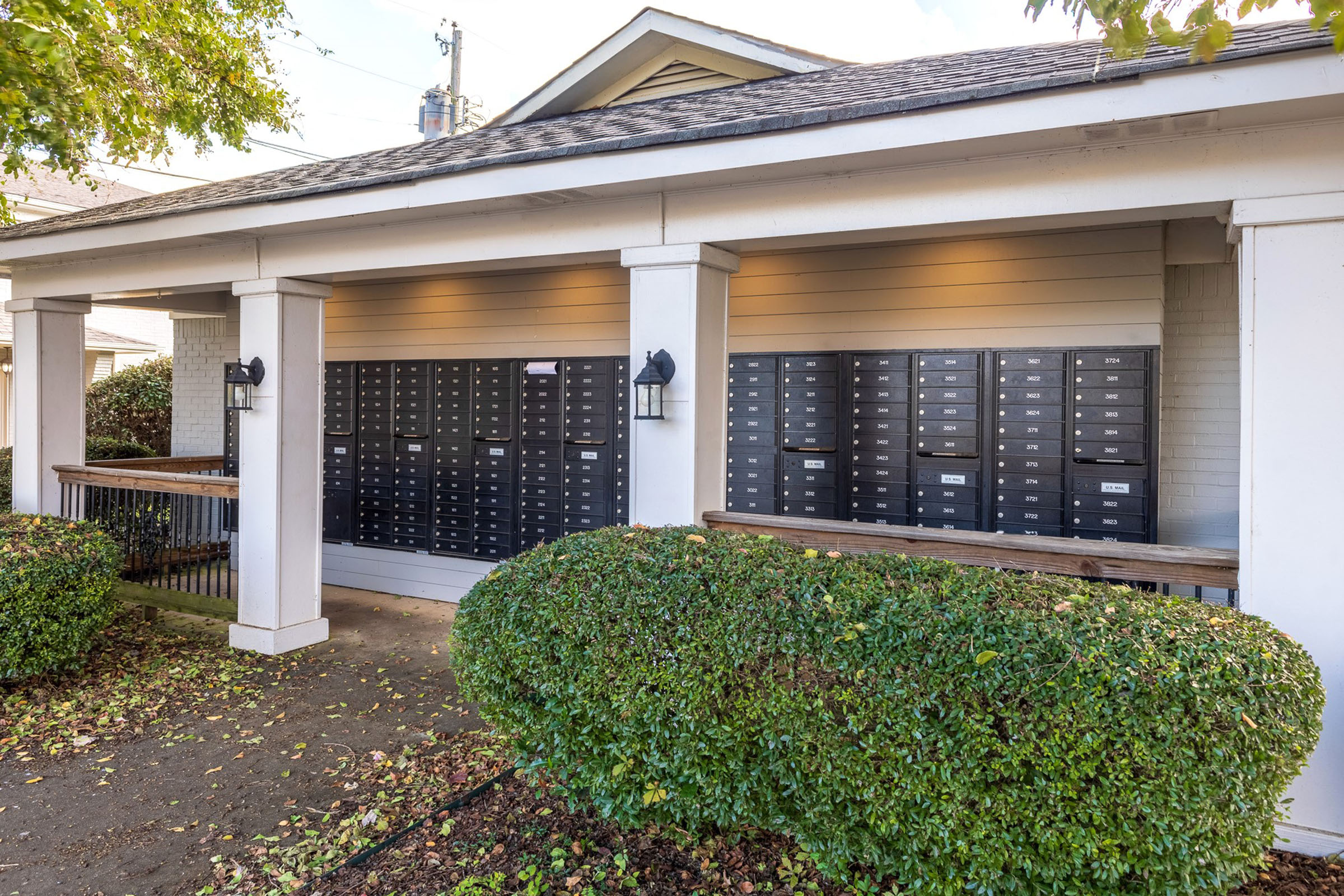 A row of black mailboxes mounted on a wall under a roof, with two white columns and outdoor lighting. The setting is surrounded by neatly trimmed greenery and fallen leaves on the ground, indicating an outdoor area near a residential building.