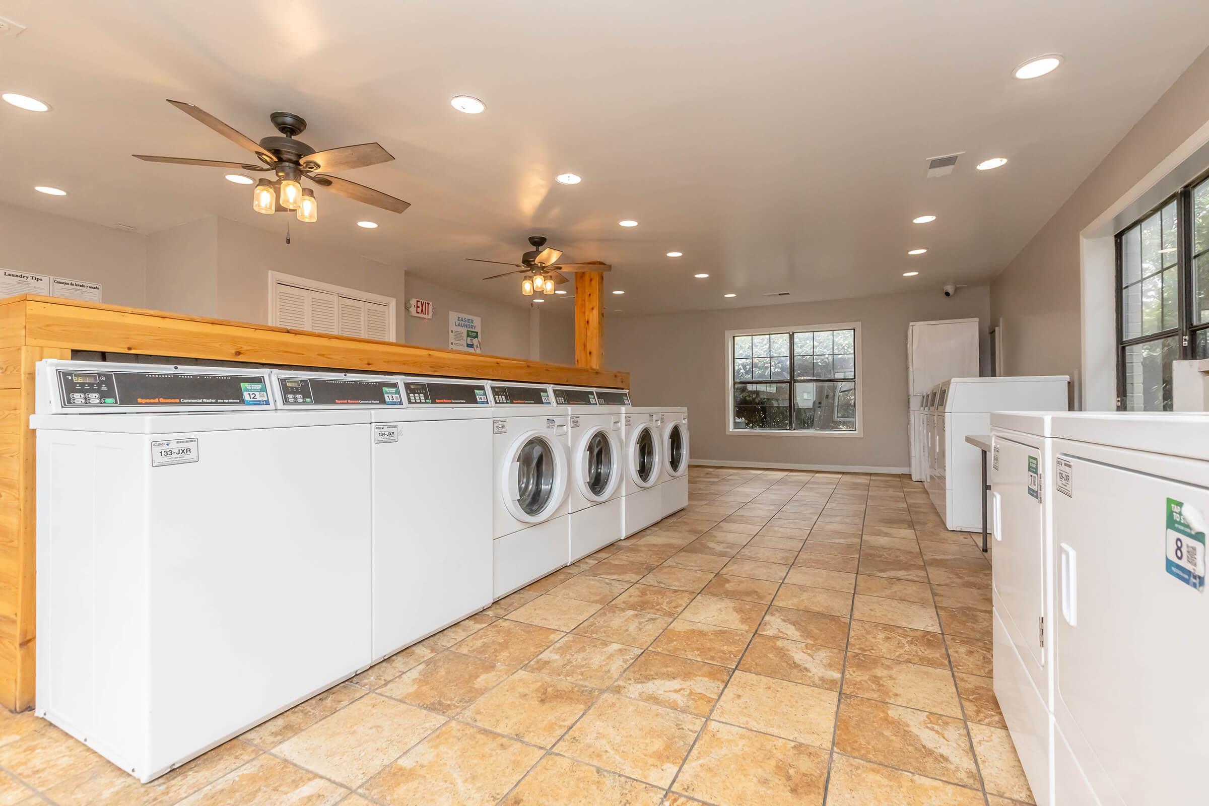 A clean, well-lit laundry room featuring multiple washing machines and dryers lined against one wall. The space has beige tiled flooring, a large window allowing natural light, and ceiling fans for ventilation. There are wooden accents and a refrigerator visible in the background.