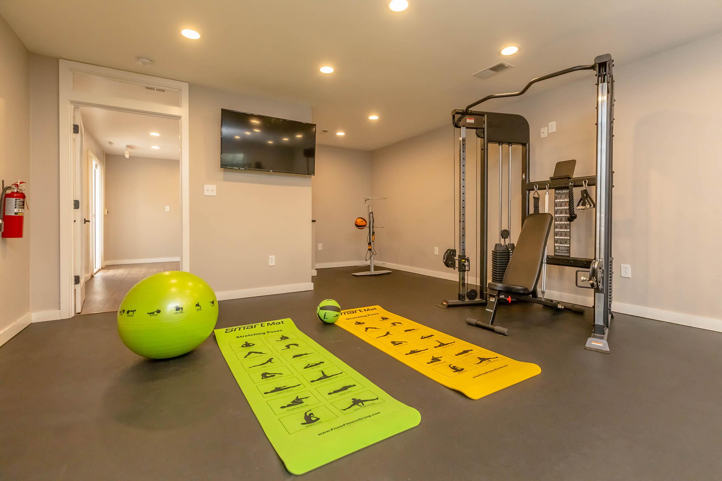A modern home gym featuring exercise mats in yellow and green, a stability ball, and a multi-gym machine. There is a wall-mounted TV and a fire extinguisher visible. The room is well-lit with overhead lights and has a neutral color scheme.