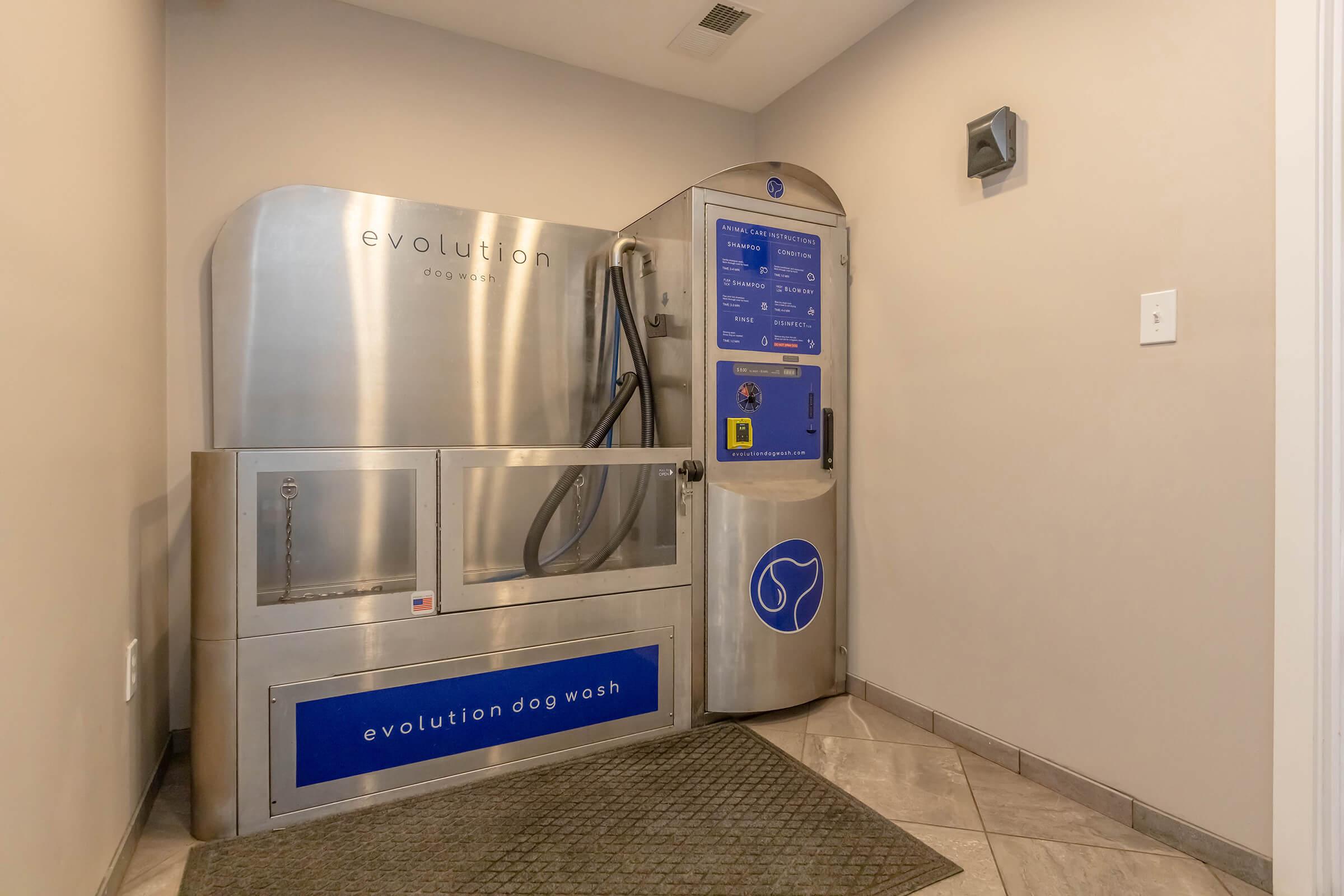 A self-service dog washing station featuring stainless steel equipment, a large wash basin, and a central control panel with instructions. The station is located in a well-lit area with tile flooring and a mat in front.