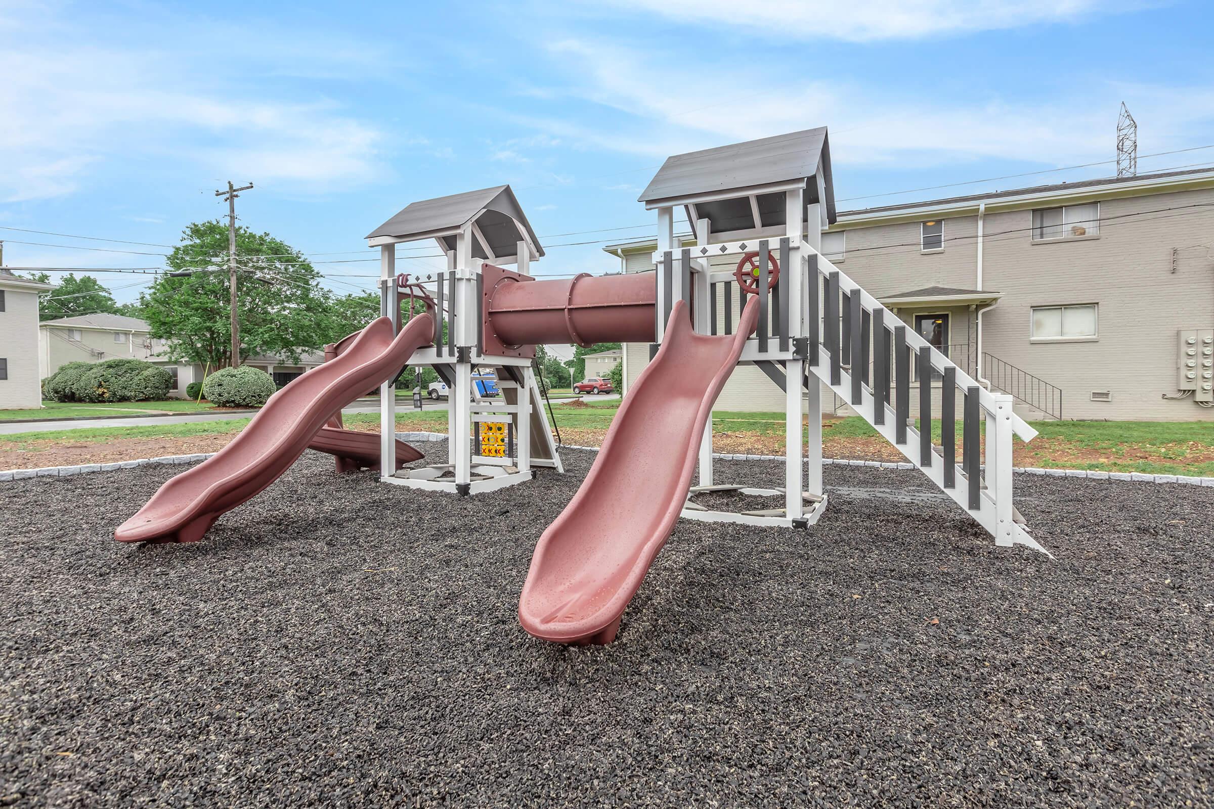 A playground structure featuring two brown slides, a central tunnel, and a staircase. It is set on a gravel surface with grass in the background, and residential buildings are visible nearby under a clear blue sky.