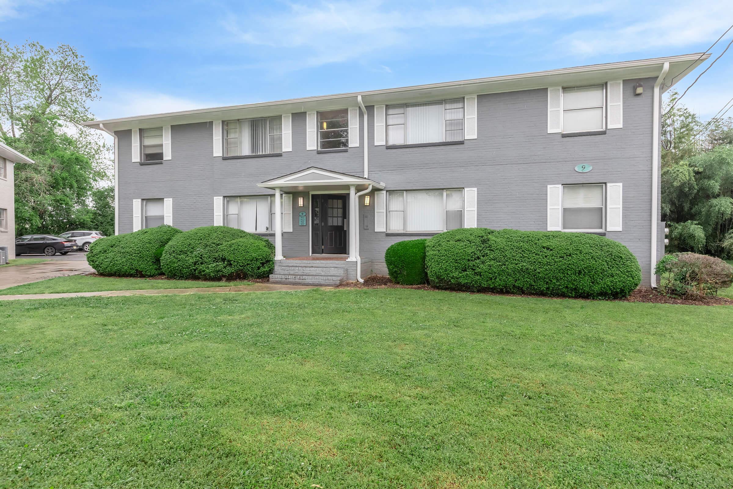 A gray two-story apartment building with white windows and a central entrance. There are neatly trimmed bushes in front and a well-maintained lawn. Trees and additional parking are visible in the background under a clear blue sky.