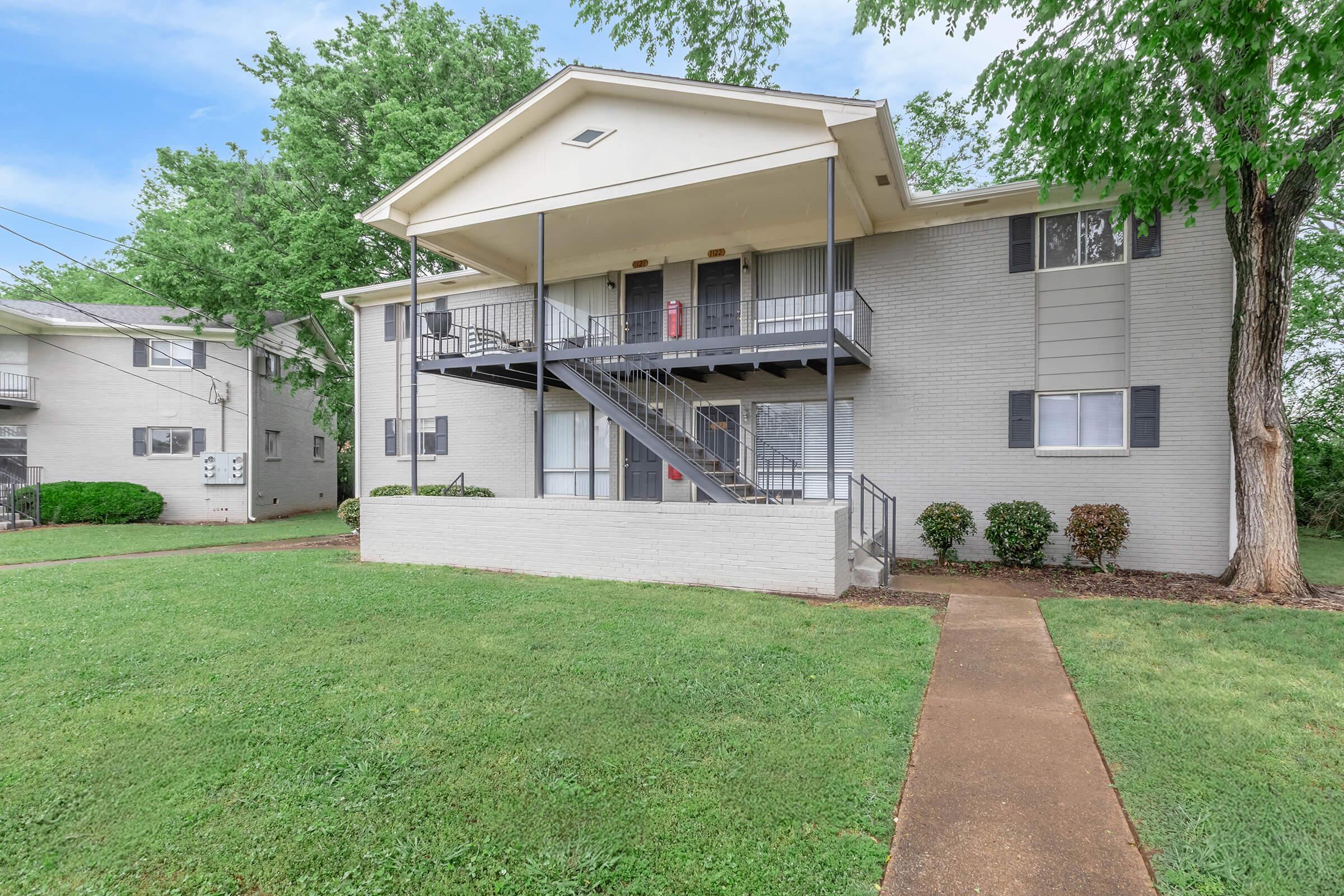 A two-story apartment building with a gray exterior, featuring a central staircase on the left side. The surrounding area includes a well-maintained lawn and small bushes. Trees provide greenery in the background, and nearby buildings are partially visible. The sky is blue with some clouds.