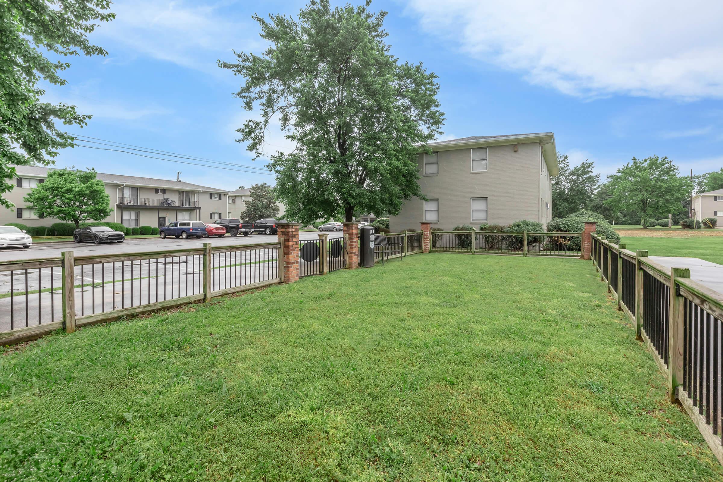 A grassy area enclosed by a wooden fence, featuring a tree and a brick pillar. In the background, there are multiple apartment buildings and parked cars visible. The sky is blue with light clouds, creating a calm, serene atmosphere.