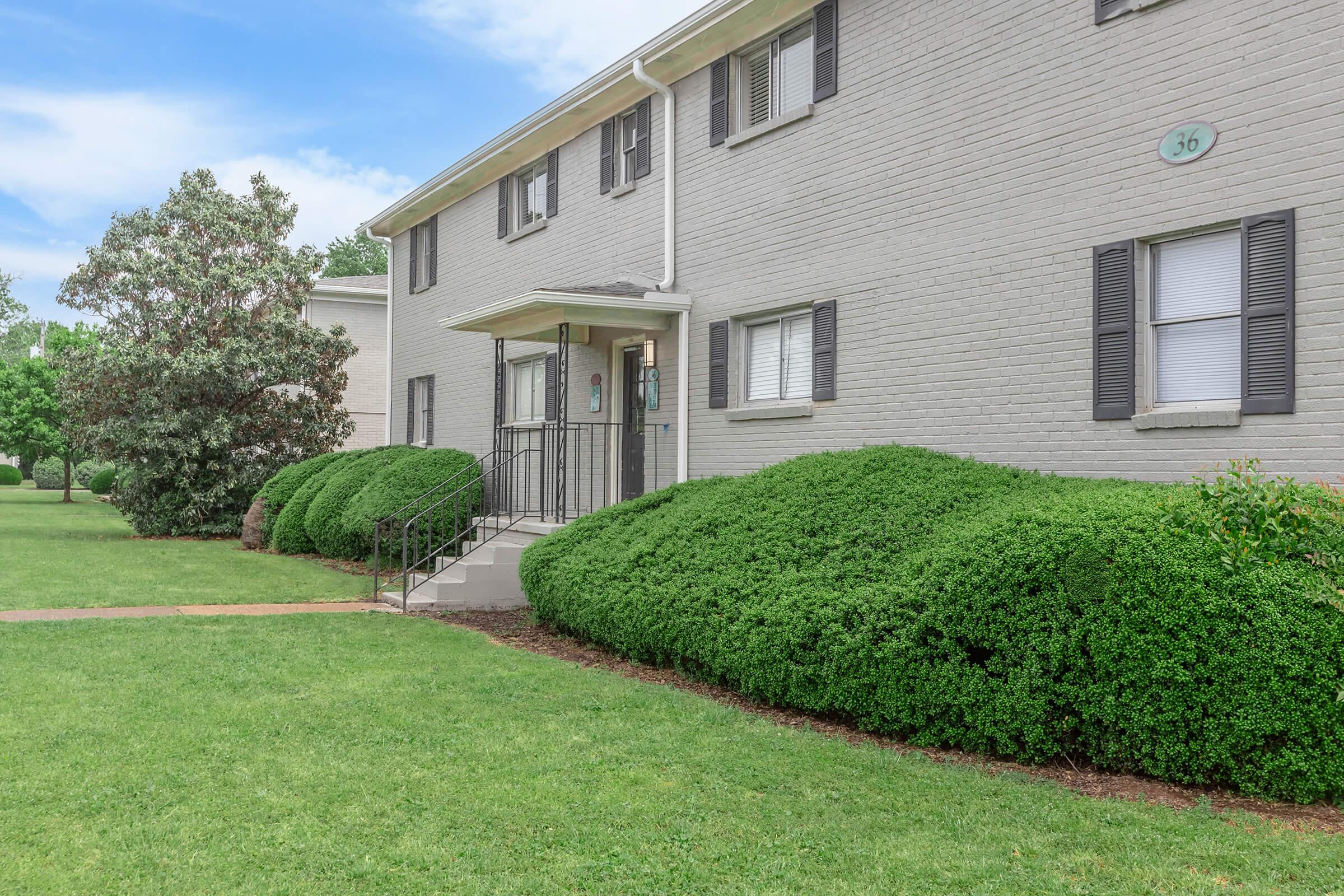 A gray apartment building with multiple windows and black shutters. A set of stairs leads to the entrance, flanked by neatly trimmed bushes. The lawn is green and well-maintained, with trees visible in the background under a clear blue sky.