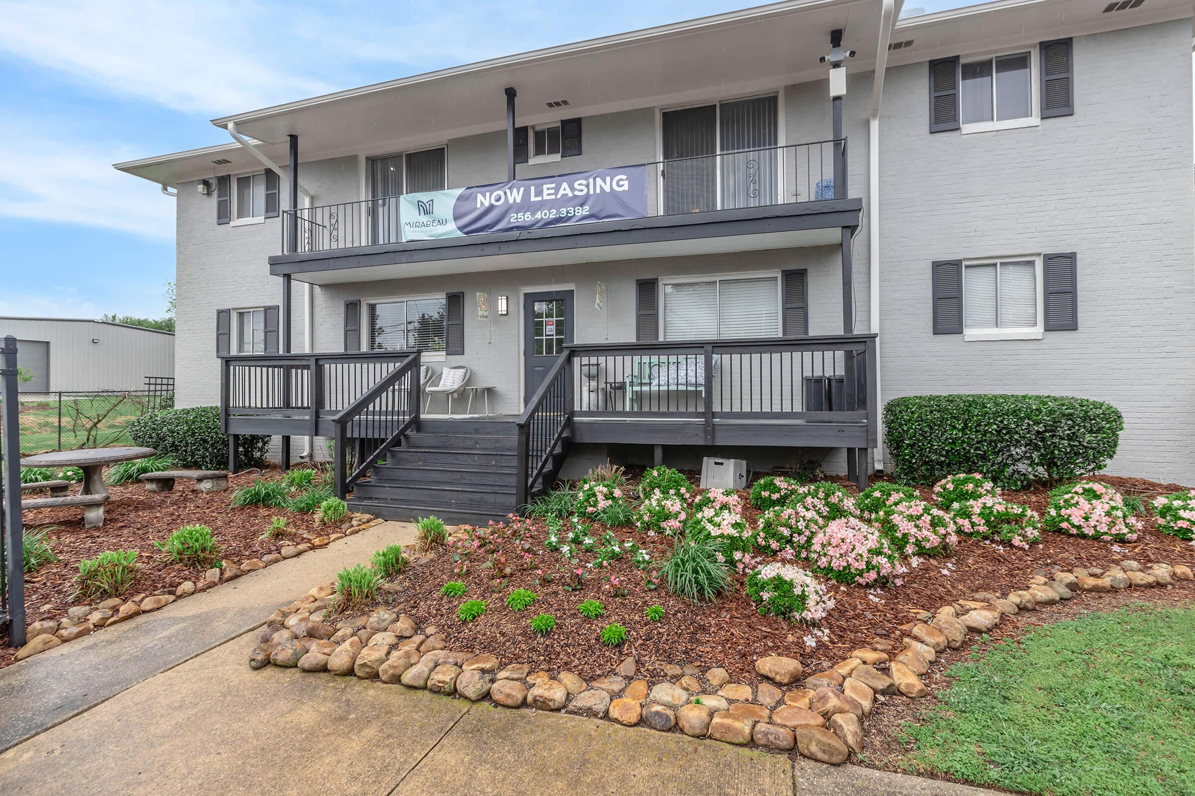 A two-story gray building with a welcoming front porch featuring a black railing. There is a "Now Leasing" banner hanging on the front, and the area is landscaped with bushes and colorful flowers. A stone pathway leads up to the entrance, providing access to outdoor seating and well-kept grounds.