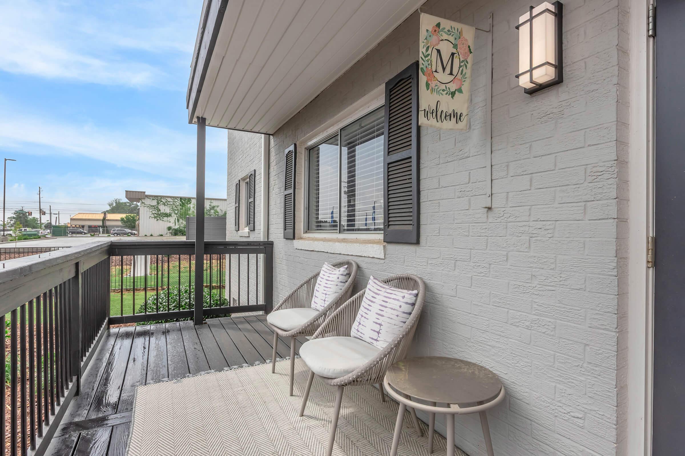A cozy outdoor space featuring two wicker chairs and a small round table on a wooden deck. There is a welcome sign hanging on the wall, and fresh greenery in the background. The area is well-lit with a wall sconce, creating an inviting atmosphere.