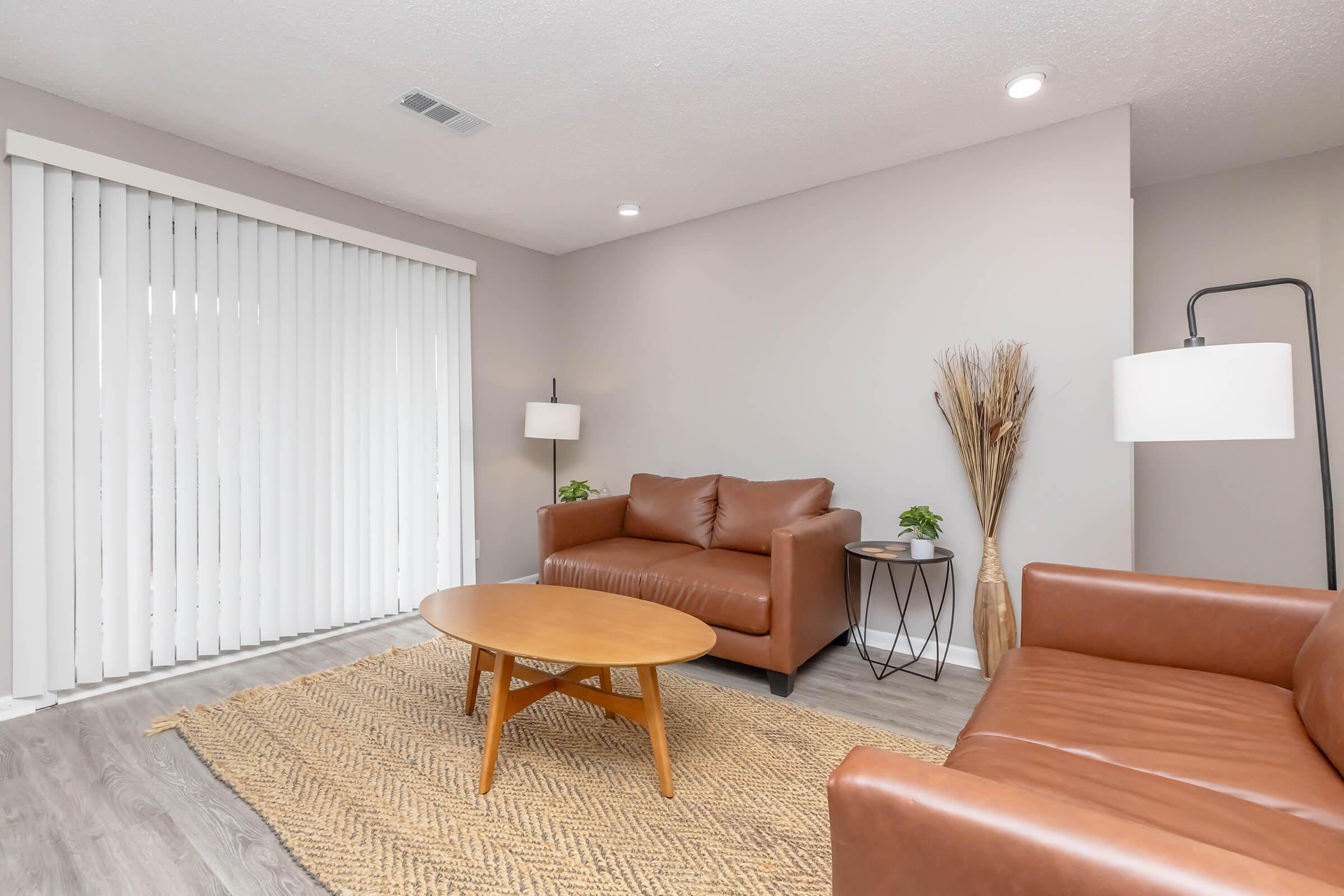 A cozy living room featuring two brown leather sofas, a round wooden coffee table, and a small side table with a plant. The space has light gray walls and wooden flooring, with a textured area rug. Large sliding glass doors are covered with white vertical blinds, allowing natural light to enter.
