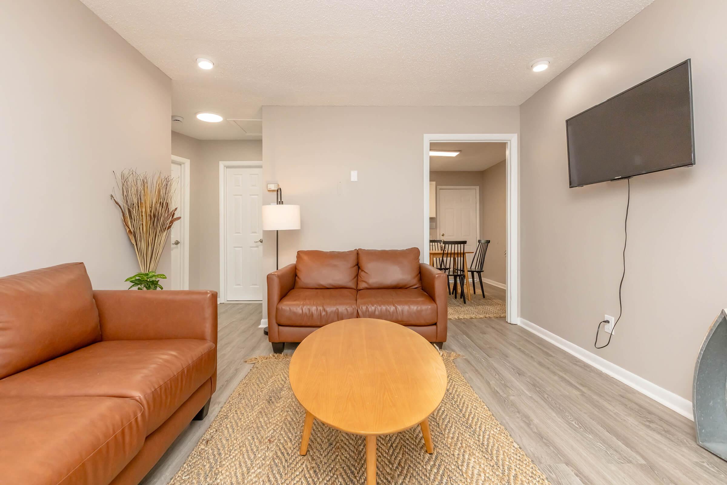 A cozy living room featuring two brown leather sofas and a round wooden coffee table on a light-colored woven rug. The walls are painted a soft gray, and there is a wall-mounted television. A doorway leads to a dining area in the background, creating a warm and inviting atmosphere.