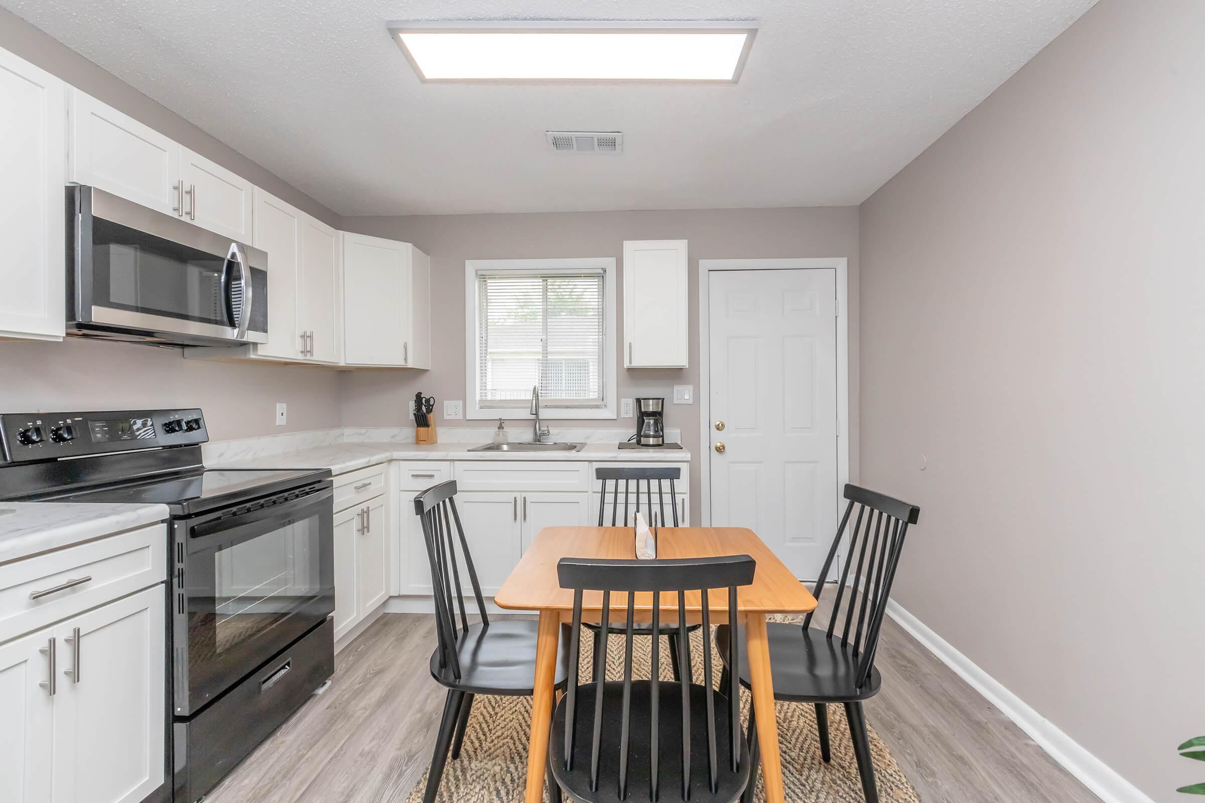 A modern kitchen featuring white cabinetry, a stainless steel microwave and oven, a sink under a window, and a small dining table with four black chairs. The flooring is a light wood look, and there is a neutral wall color, creating a bright and inviting atmosphere.