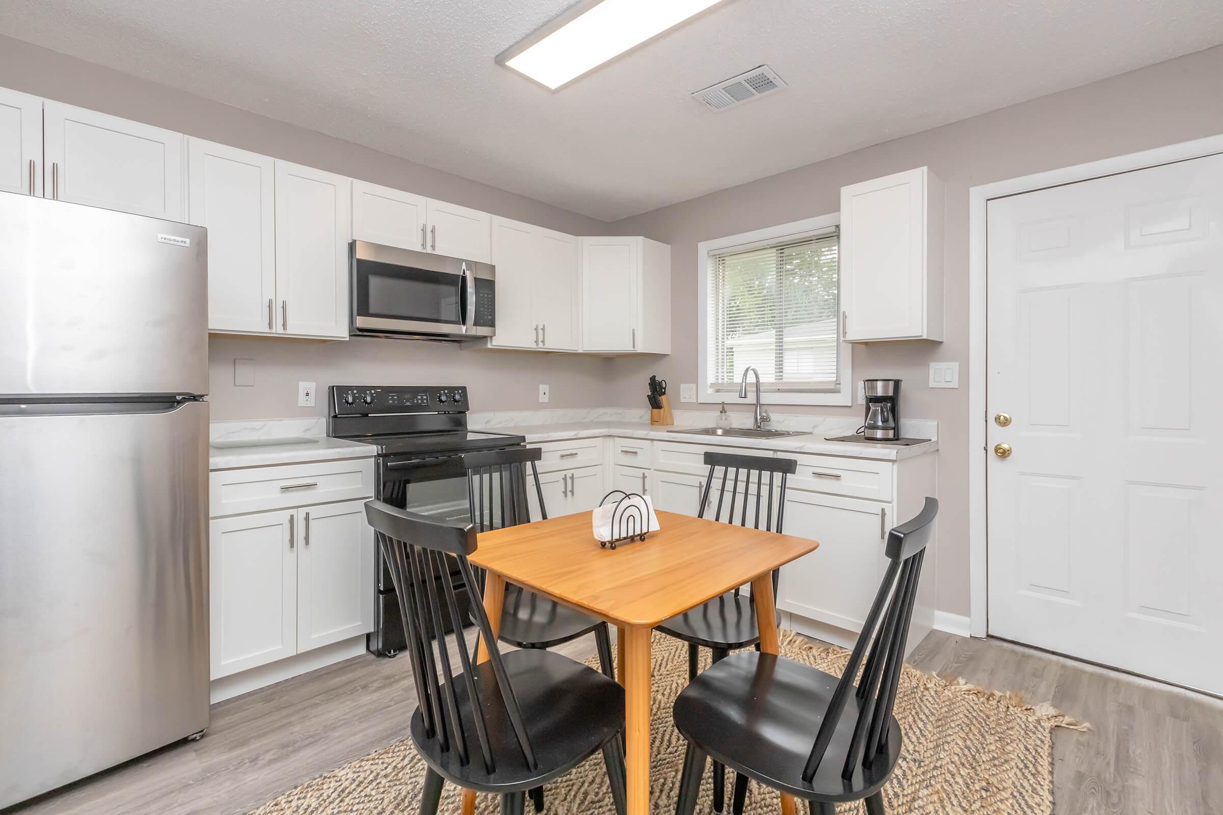 A modern kitchen featuring white cabinetry, stainless steel appliances, and a wood dining table with four black chairs. The countertop is decorated with a small centerpiece, and a door leads to the outside. Natural light enters through a window, enhancing the inviting atmosphere.