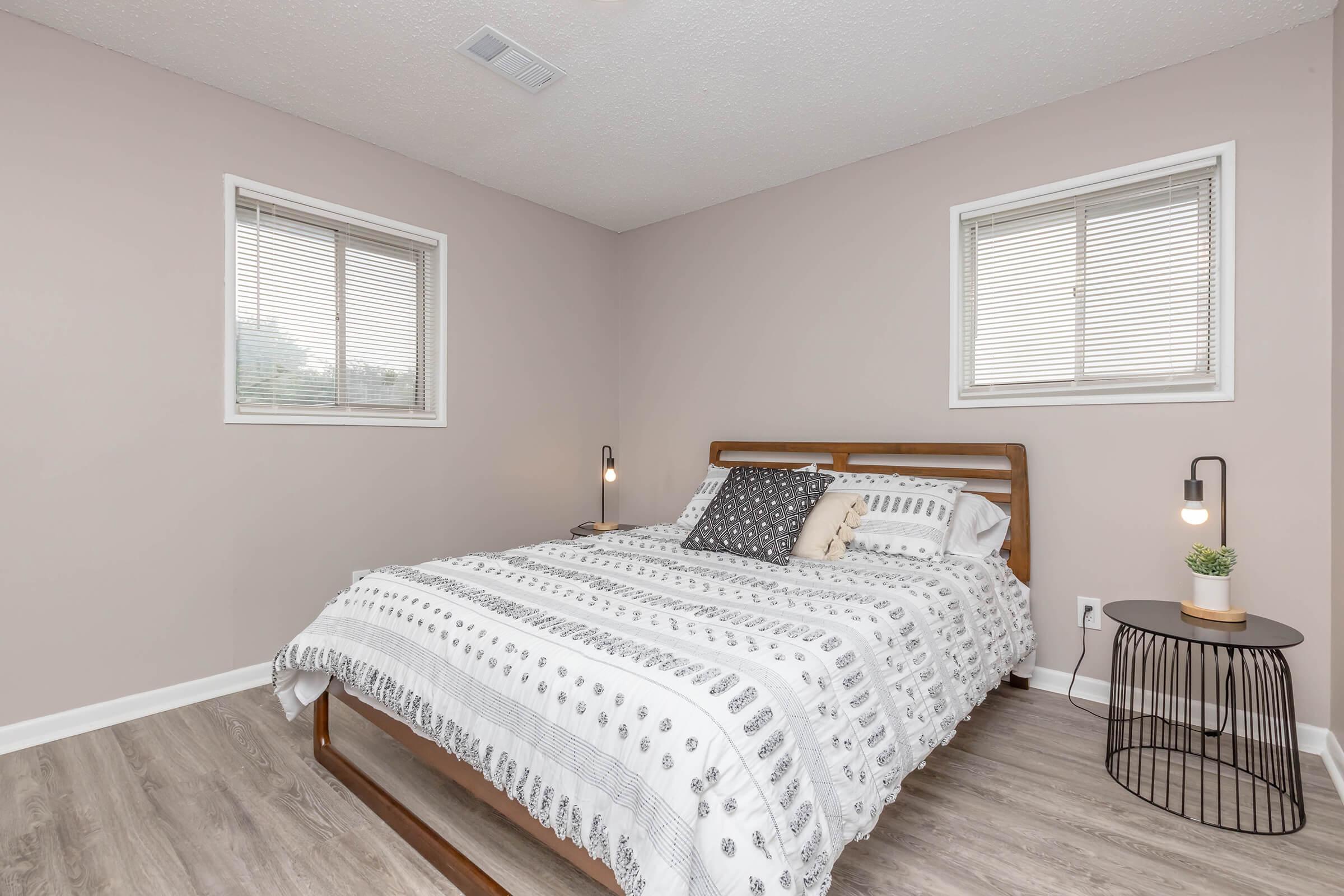A neatly designed bedroom featuring a wooden bed frame with a patterned blanket, two decorative pillows, and a bedside table. Natural light filters through two windows with blinds, and the walls are painted a soft pink color. The wooden floor adds warmth to the space.