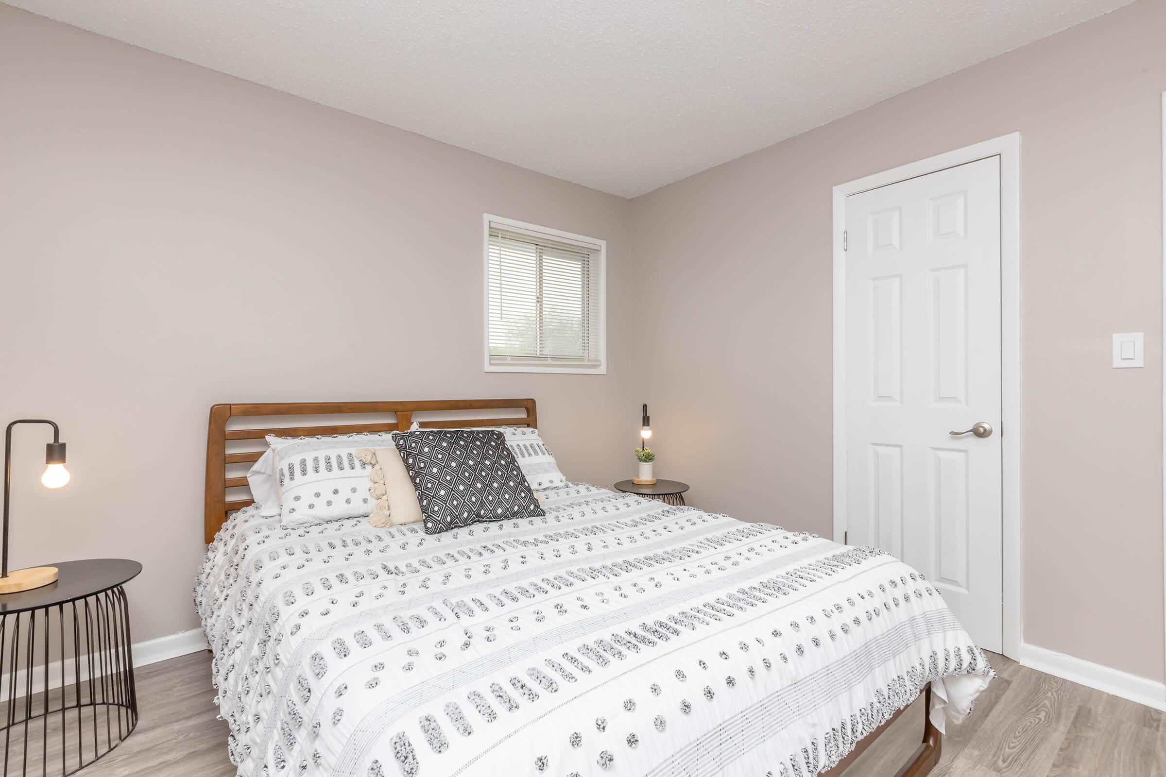A cozy bedroom featuring a wooden bed with a striped white and gray duvet cover, accent pillows, a small nightstand with a lamp, and a decorative plant. The walls are painted in a soft beige tone, with a window allowing natural light to brighten the space. A door leads to another area.