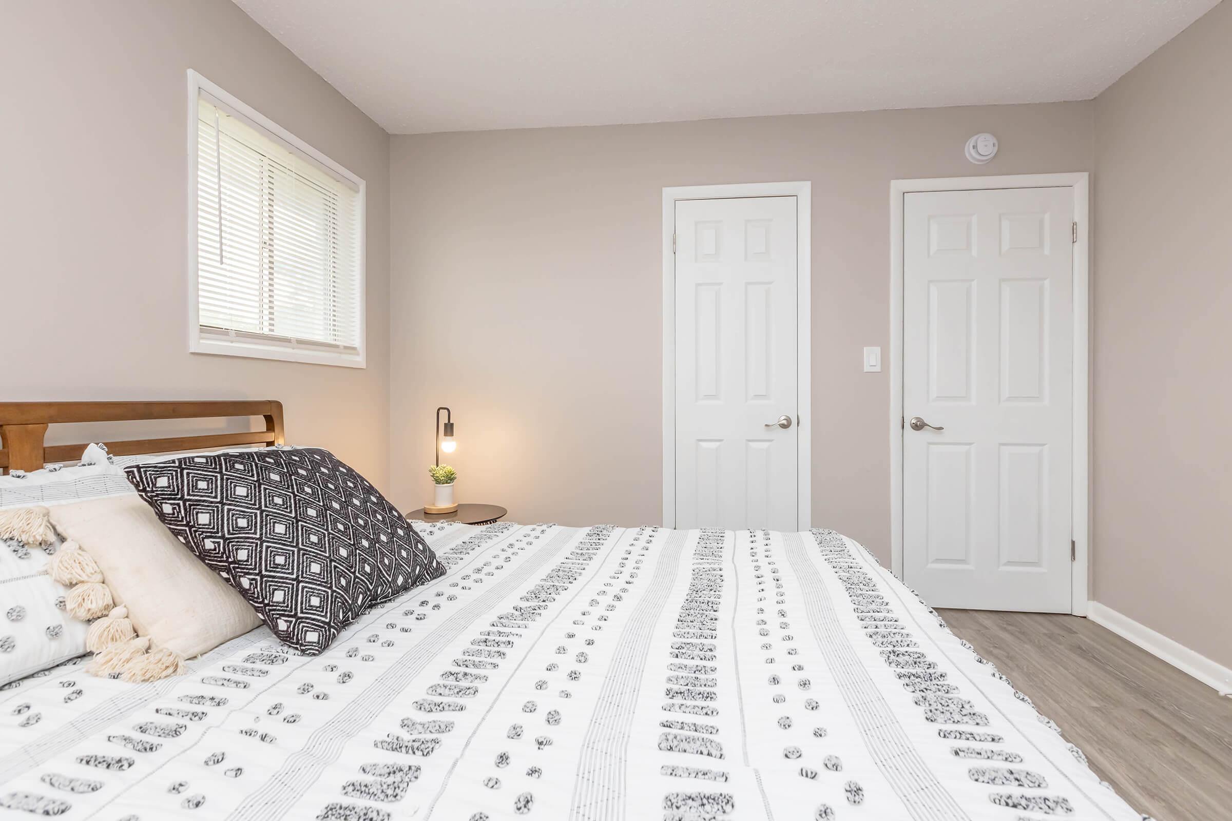 A cozy bedroom featuring a wooden bed with patterned bedding, two closed white doors on the right, and a small lamp on a bedside table. The walls are painted a soft beige color and there is a window with blinds allowing natural light to enter.