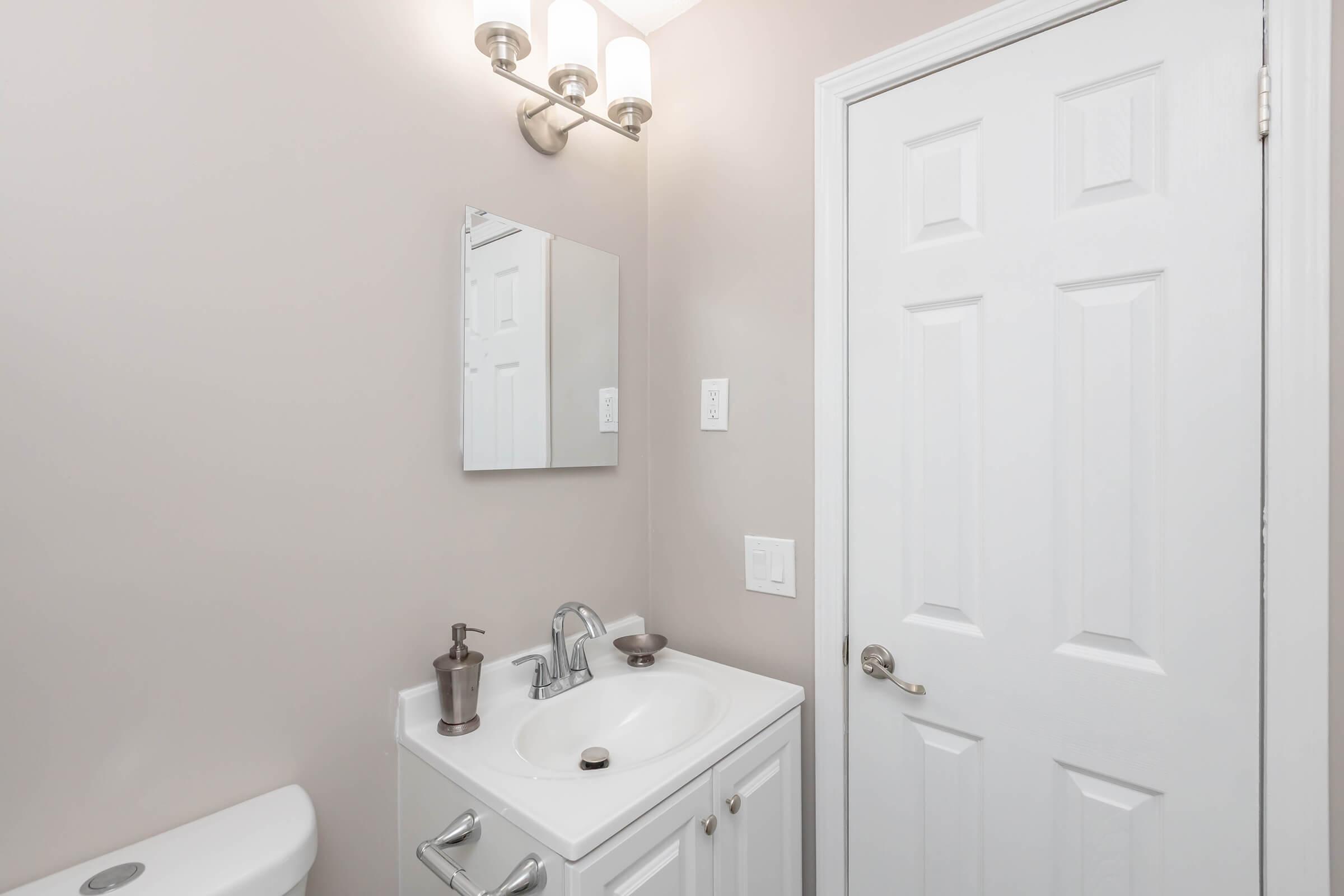 A small bathroom featuring a white vanity with a sink, a wall-mounted mirror above it, a light fixture with three bulbs, a white door leading to another room, and a toilet. The walls are painted in a light shade, creating a clean and modern look.