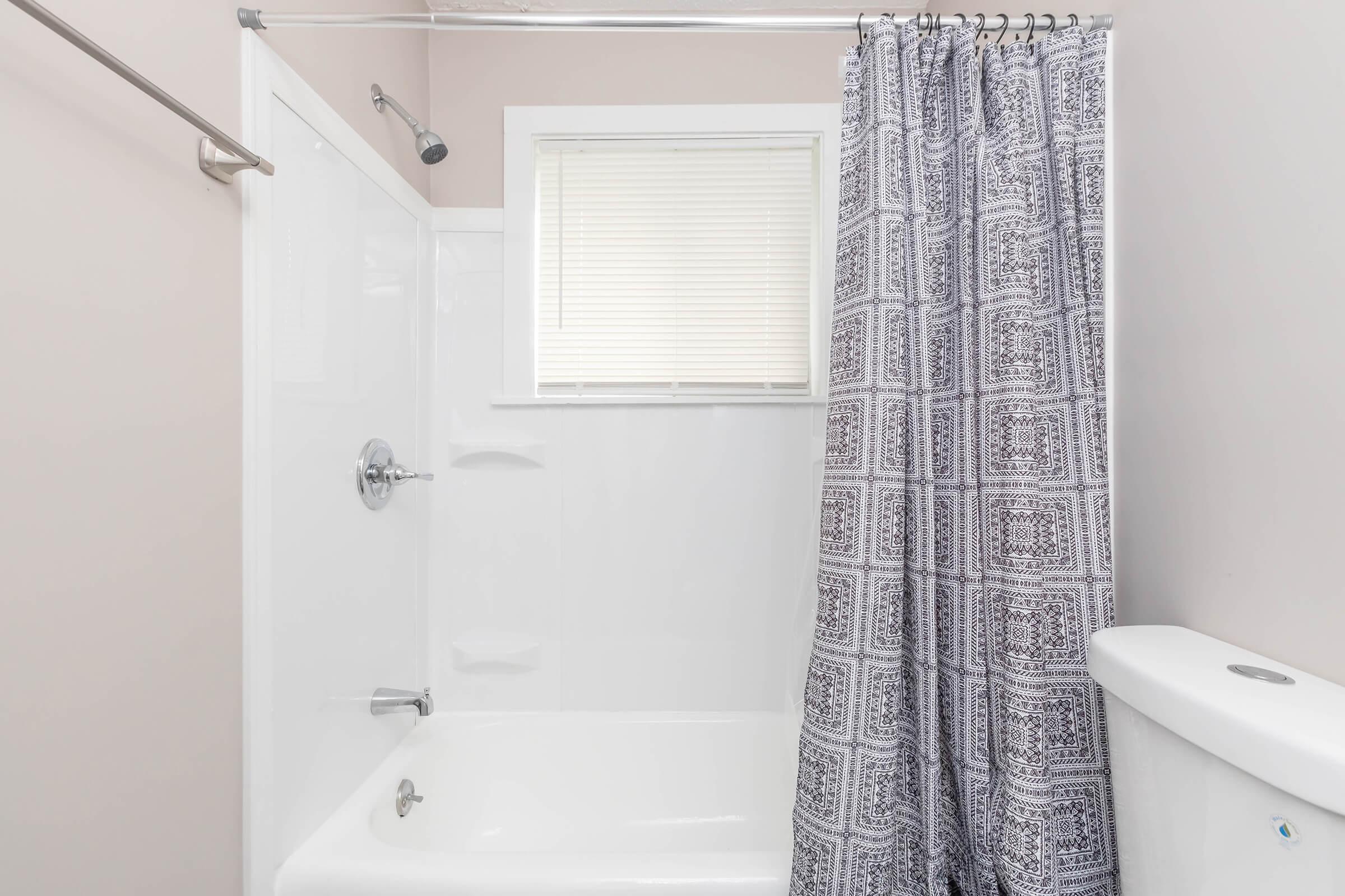A clean and modern bathroom featuring a white tub and shower with a patterned gray curtain. The walls are painted a soft pink, and there is a window with blinds allowing natural light. A white toilet is visible beside the tub.