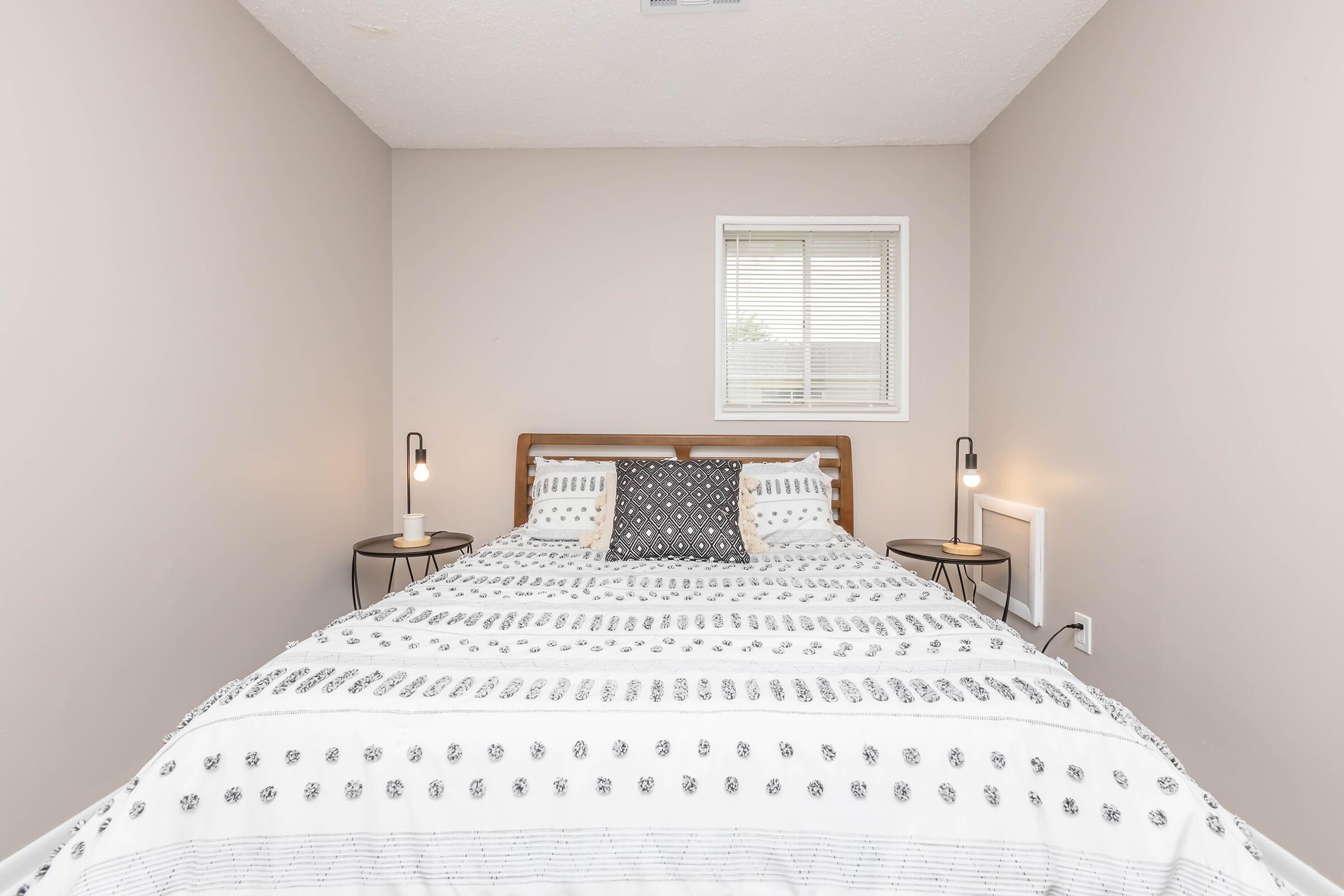 A cozy bedroom featuring a wooden bed with a white and black patterned comforter. There are two bedside tables with modern lamps, and a small window letting in natural light. The walls are painted a light beige, creating a warm and inviting atmosphere.