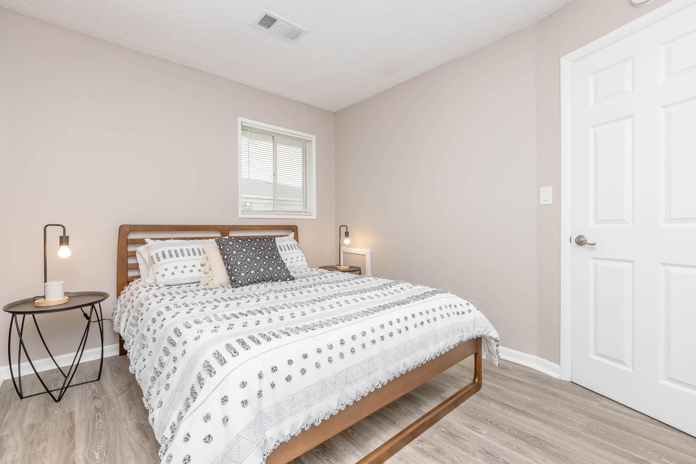 A cozy bedroom featuring a wooden bed with a white and gray striped comforter, accented by decorative pillows. There’s a bedside table with a lamp on one side and a small window allowing natural light. The walls are painted in a soft beige, and the flooring is light-colored wood. A closed white door is visible.