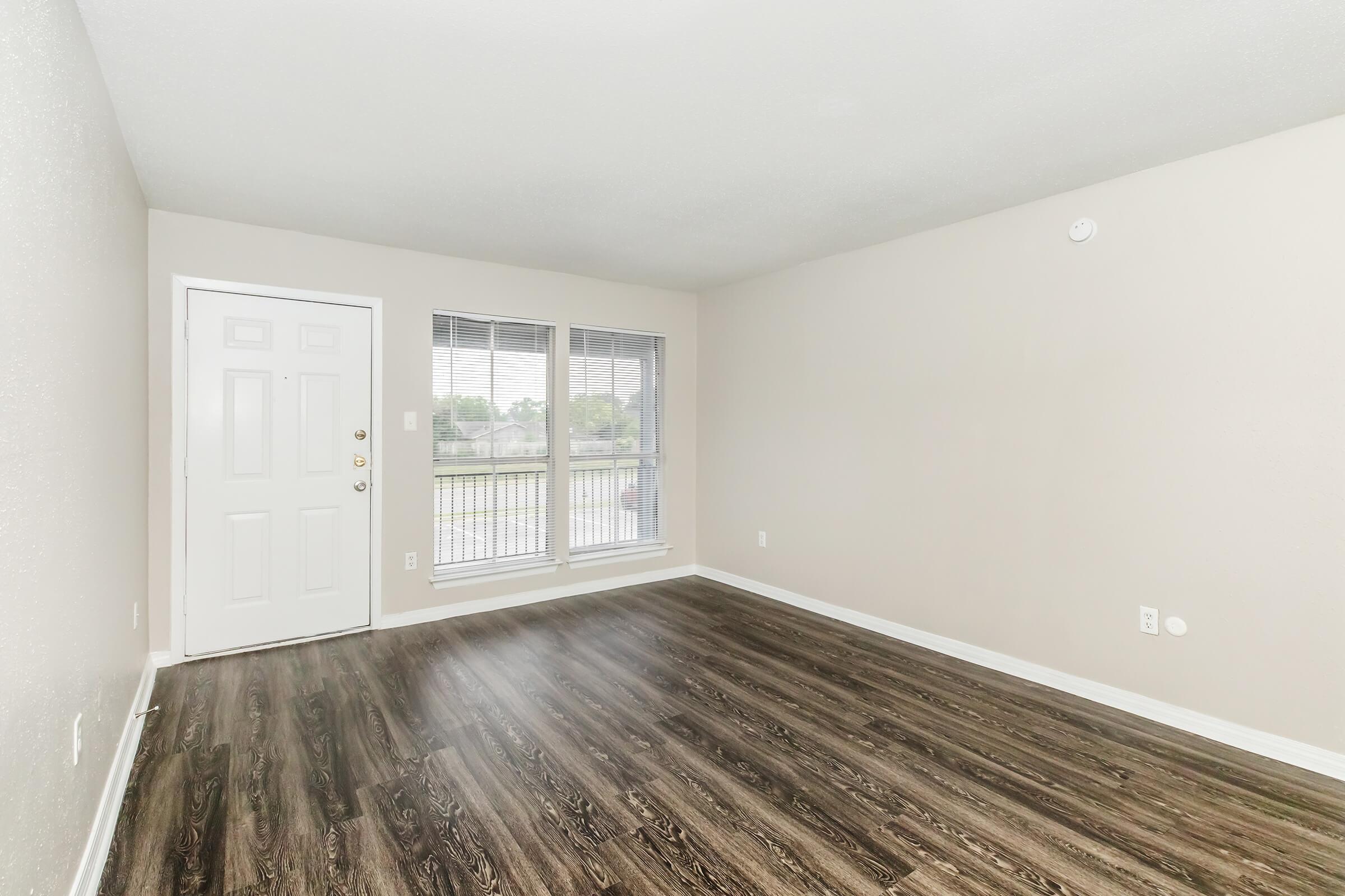A spacious, empty living room with light gray walls and dark wood-style flooring. It features a front door on the left and large windows allowing natural light to enter, offering a view of the outdoors. The room is unadorned, ideal for personalized decoration and furnishing.