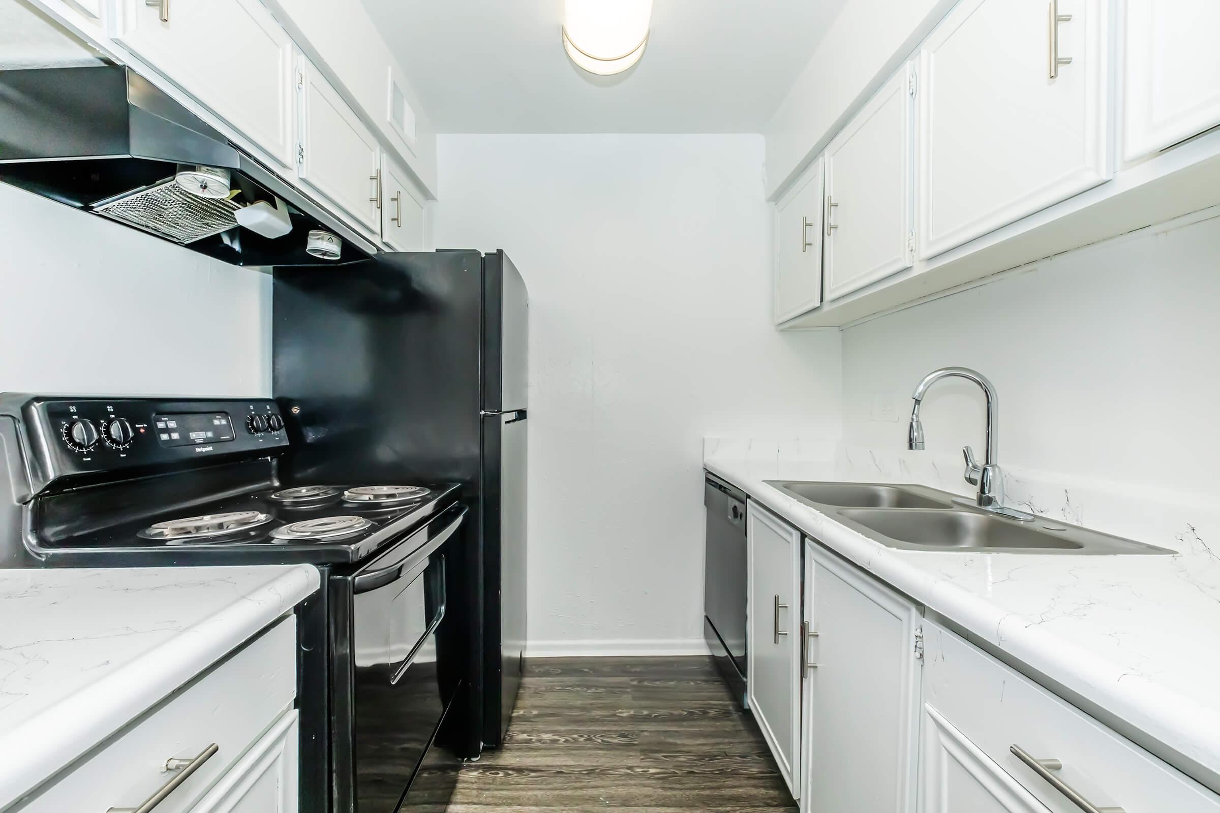A modern kitchen featuring white cabinets, a black refrigerator, an electric stove, and a stainless steel sink with a double basin. The countertops have a marble-like appearance, and the flooring is dark wood. The walls are painted white, creating a bright and clean atmosphere.