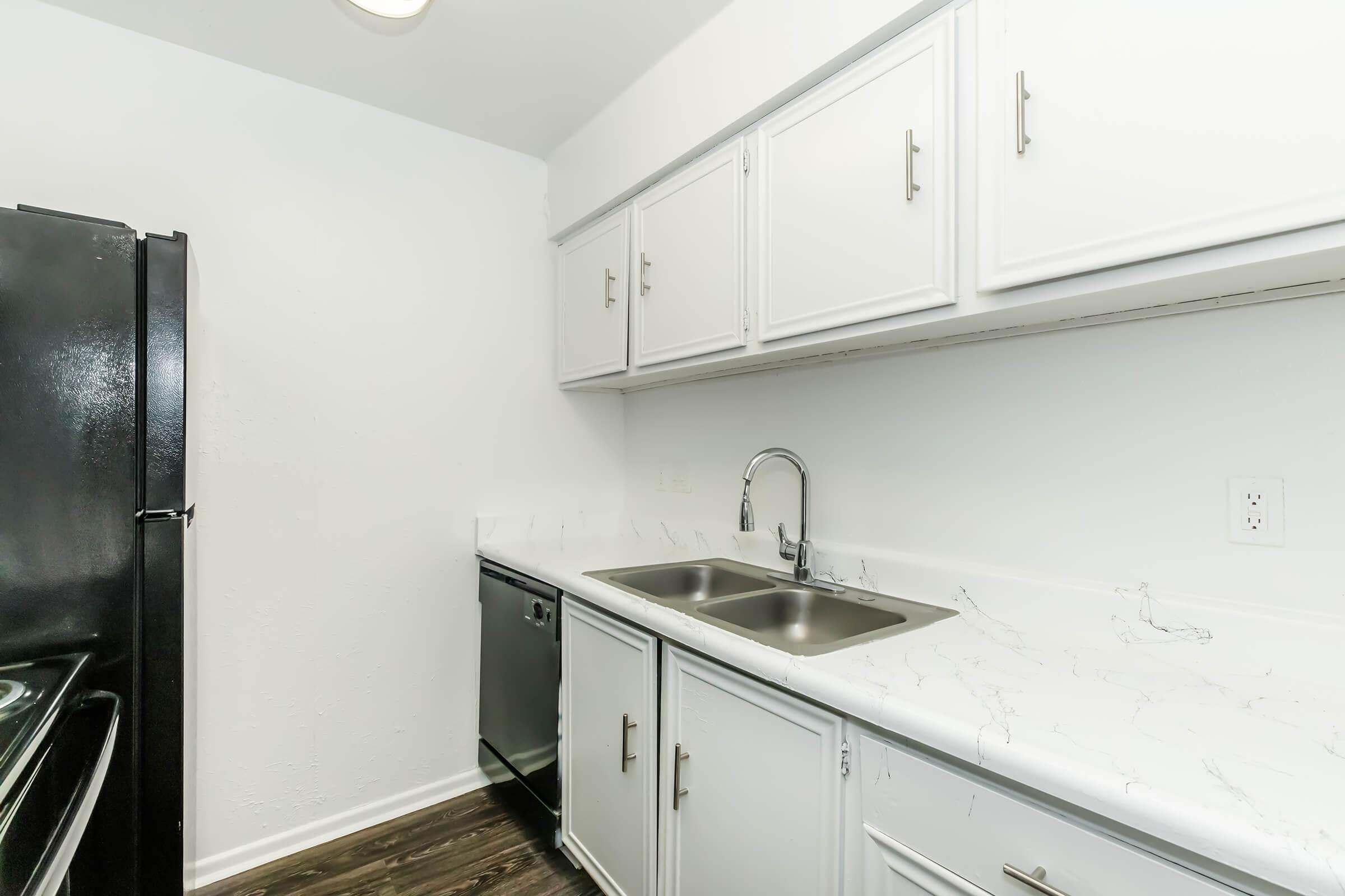 Modern kitchen featuring white cabinets, a double sink with a sleek faucet, and dark appliances including a black refrigerator and dishwasher. The countertop has a light marble-like finish, and the floor is a dark wood laminate. Walls are painted white, creating a clean and contemporary look.