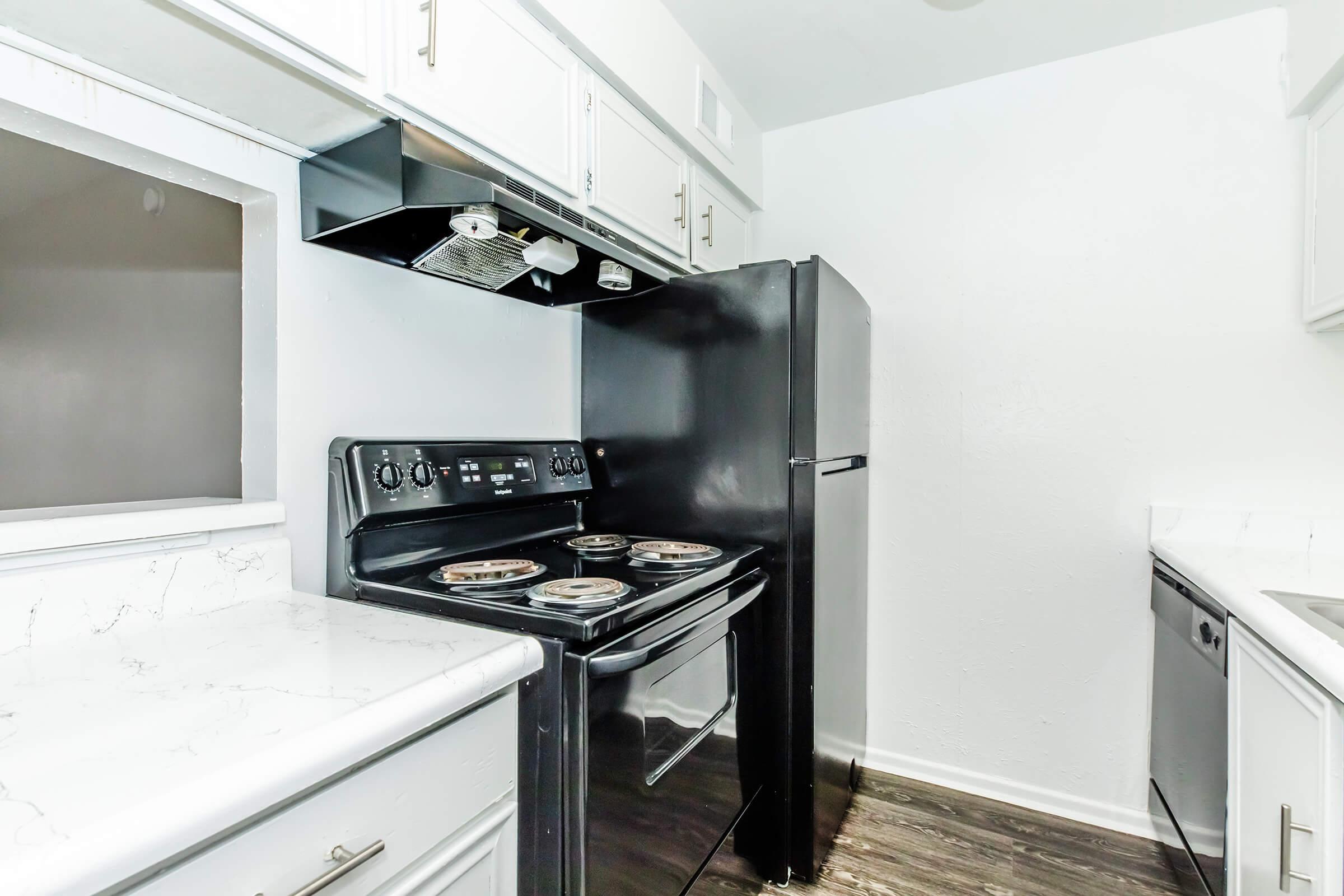 A modern kitchen featuring a black oven and refrigerator, white cabinets, and a sleek countertop. The stove has four burners and an overhead exhaust fan. The kitchen has a bright, clean aesthetic with a minimalist design and dark flooring, enhancing the overall contemporary look.