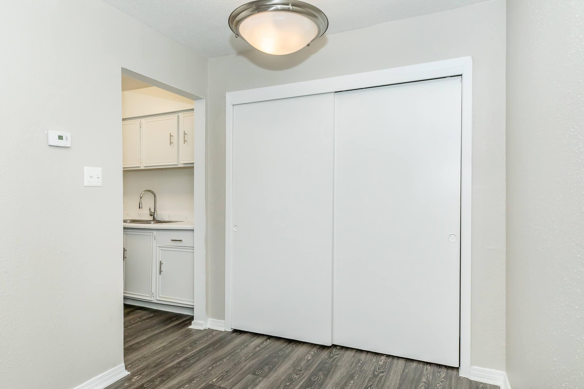 A view of a room featuring a white double sliding closet door, light-colored walls, and wood-style flooring. An adjacent area shows a glimpse of a kitchen with white cabinets and a sink. The space is well-lit by a contemporary ceiling light fixture.